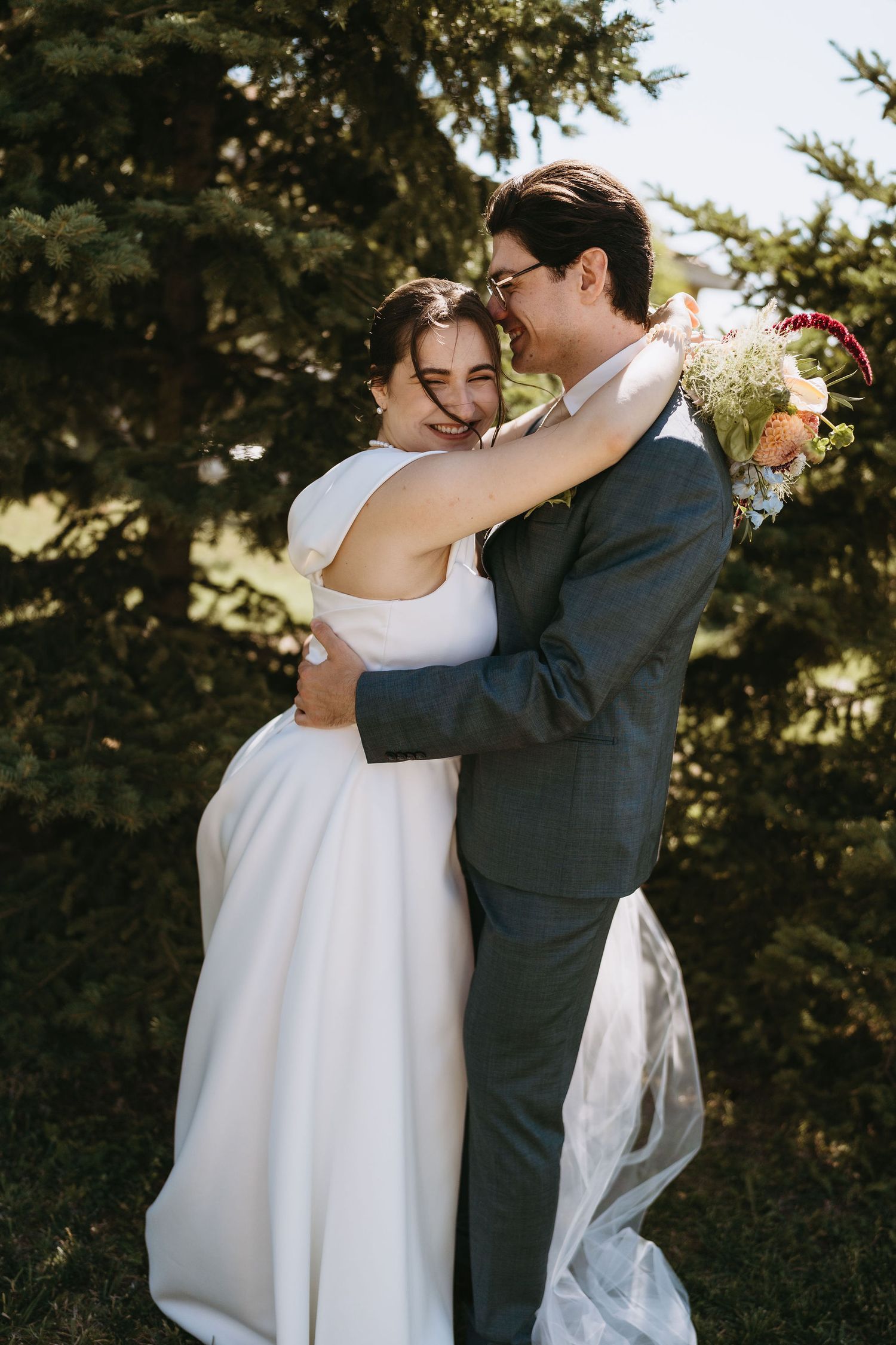 Bride and groom embracing outdoors, smiling joyfully, holding a bouquet, with evergreen trees in the background.