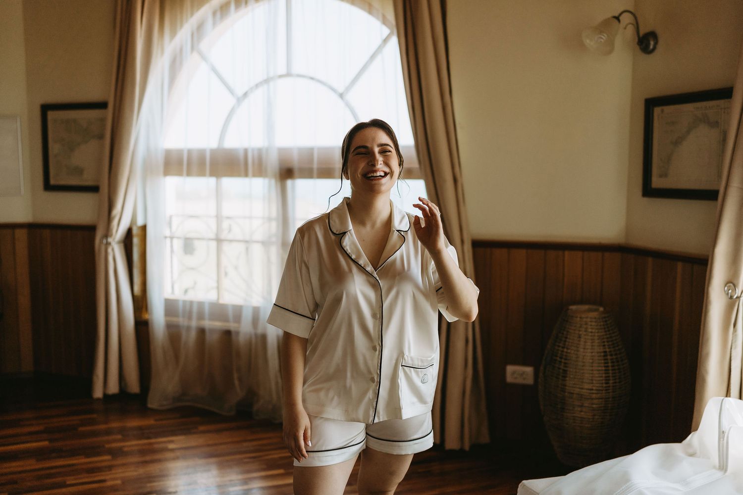 Woman in white pajamas smiling and holding earrings in a warmly lit bedroom with wooden floors.