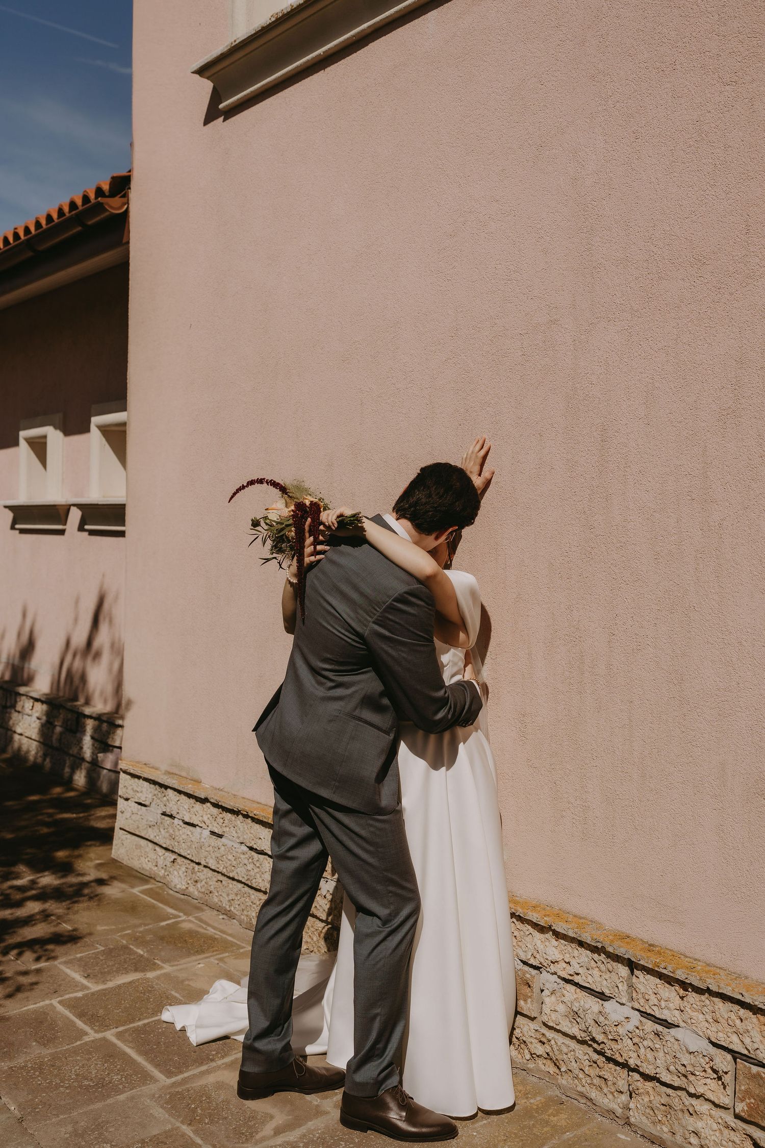 Bride and groom share tender embrace against pink stucco wall, bride holding bouquet in white gown, groom in gray suit.