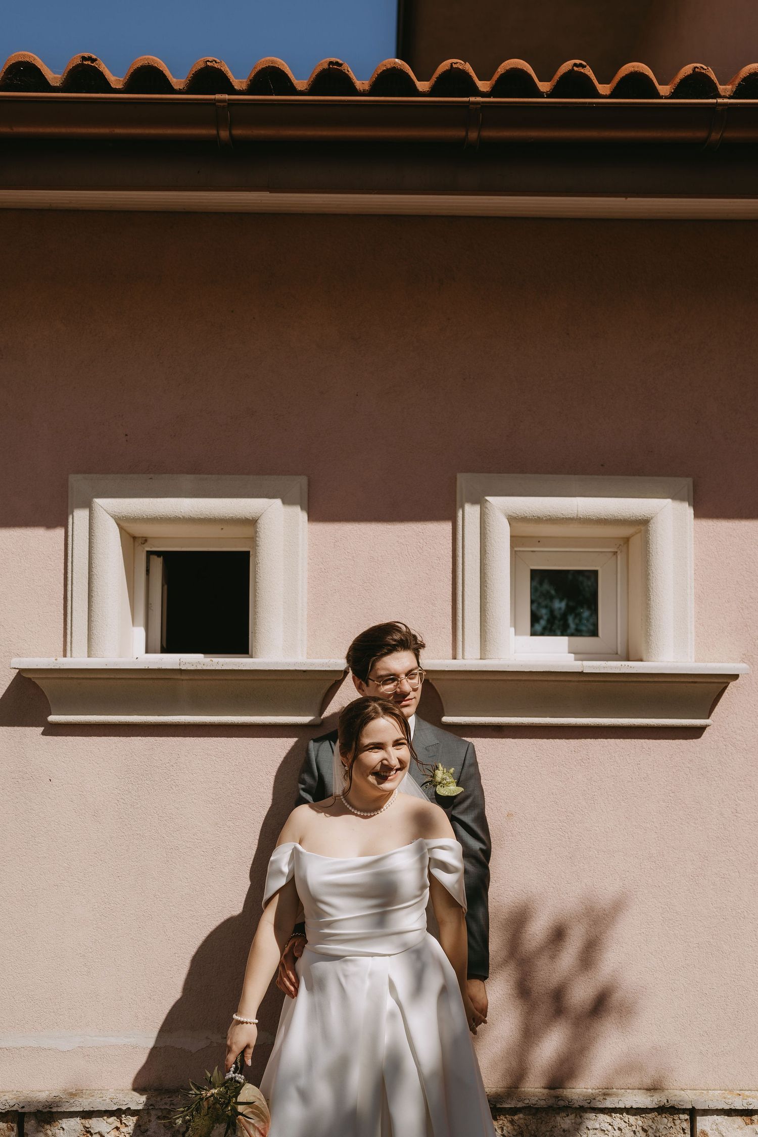 Smiling bride in white satin gown with groom behind her, posing against a pink Mediterranean villa wall.