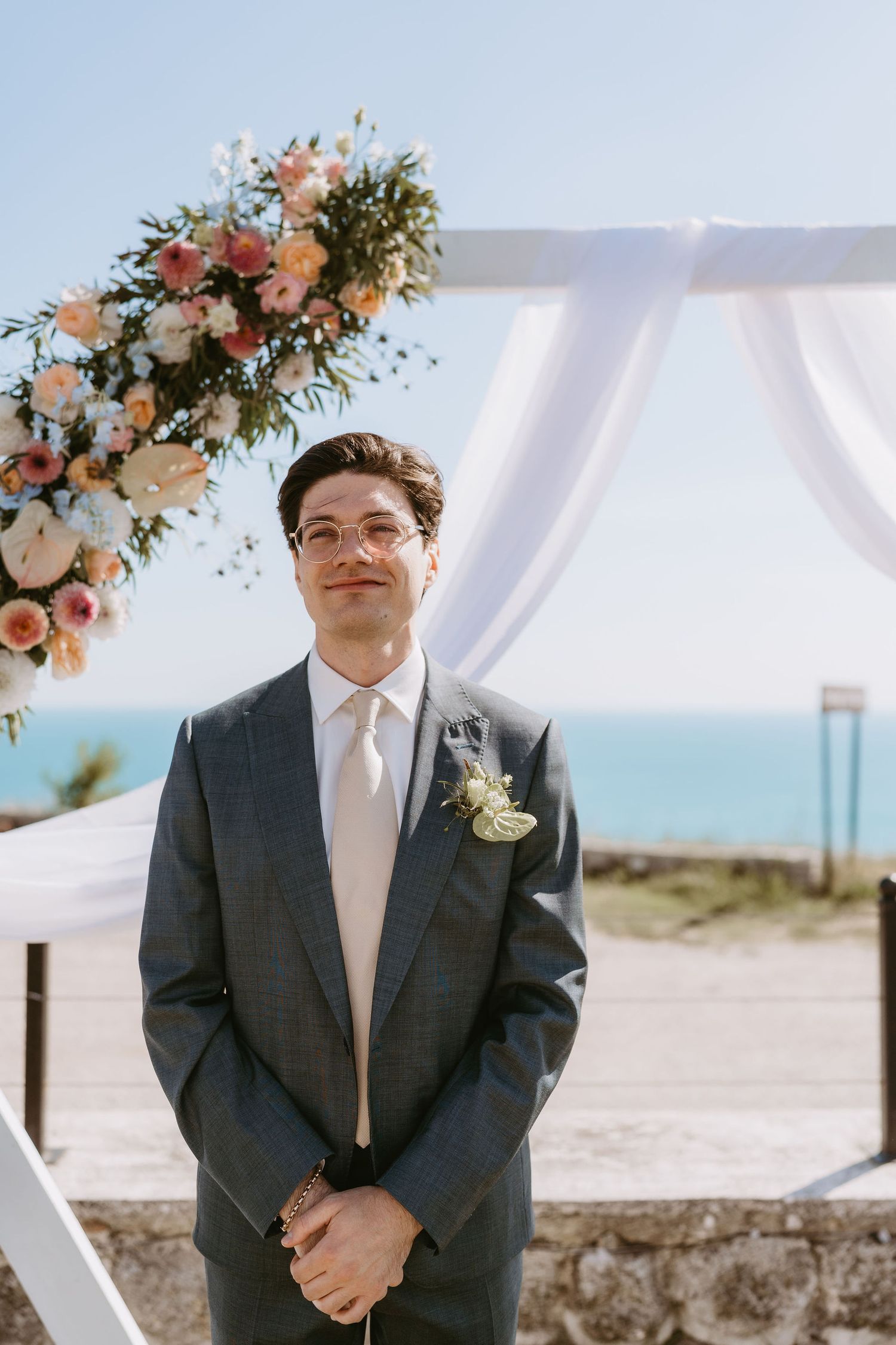 Groom in gray suit with boutonniere stands before floral arch with ocean view at outdoor wedding ceremony.