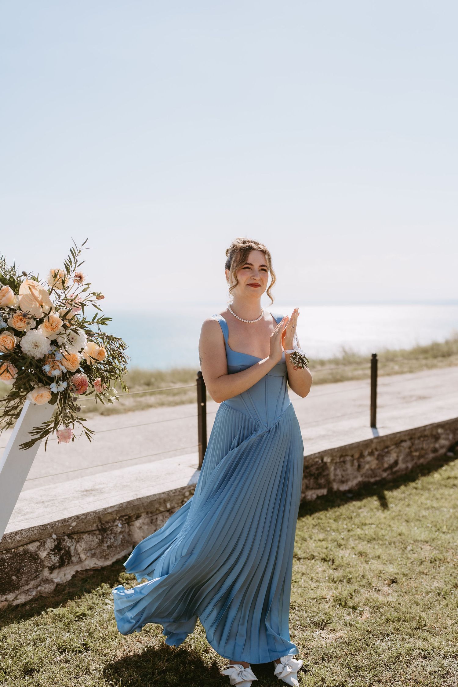 Woman in blue pleated maxi dress standing near floral arrangement with ocean view backdrop on sunny day.