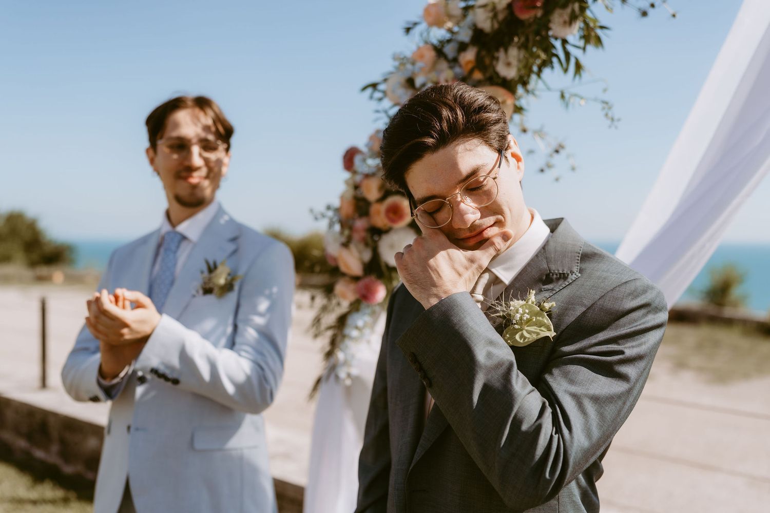 Emotional groom wiping tears at outdoor wedding ceremony with floral arch and best man clapping in background.