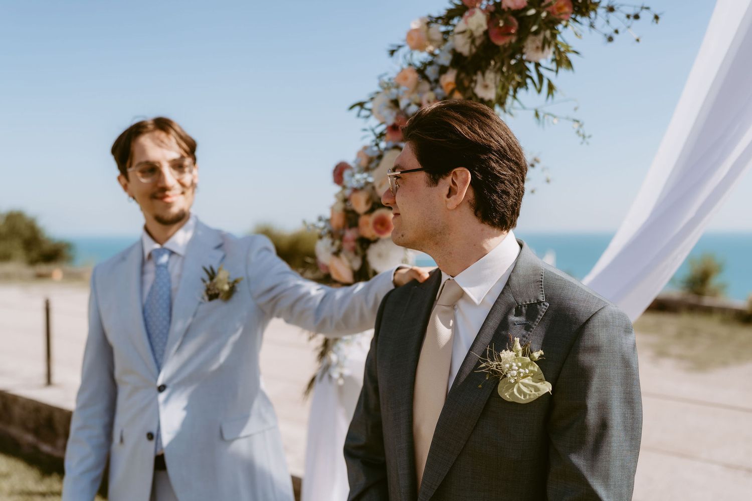 Two grooms in suits share a tender moment at an outdoor beach wedding ceremony with floral arch backdrop.