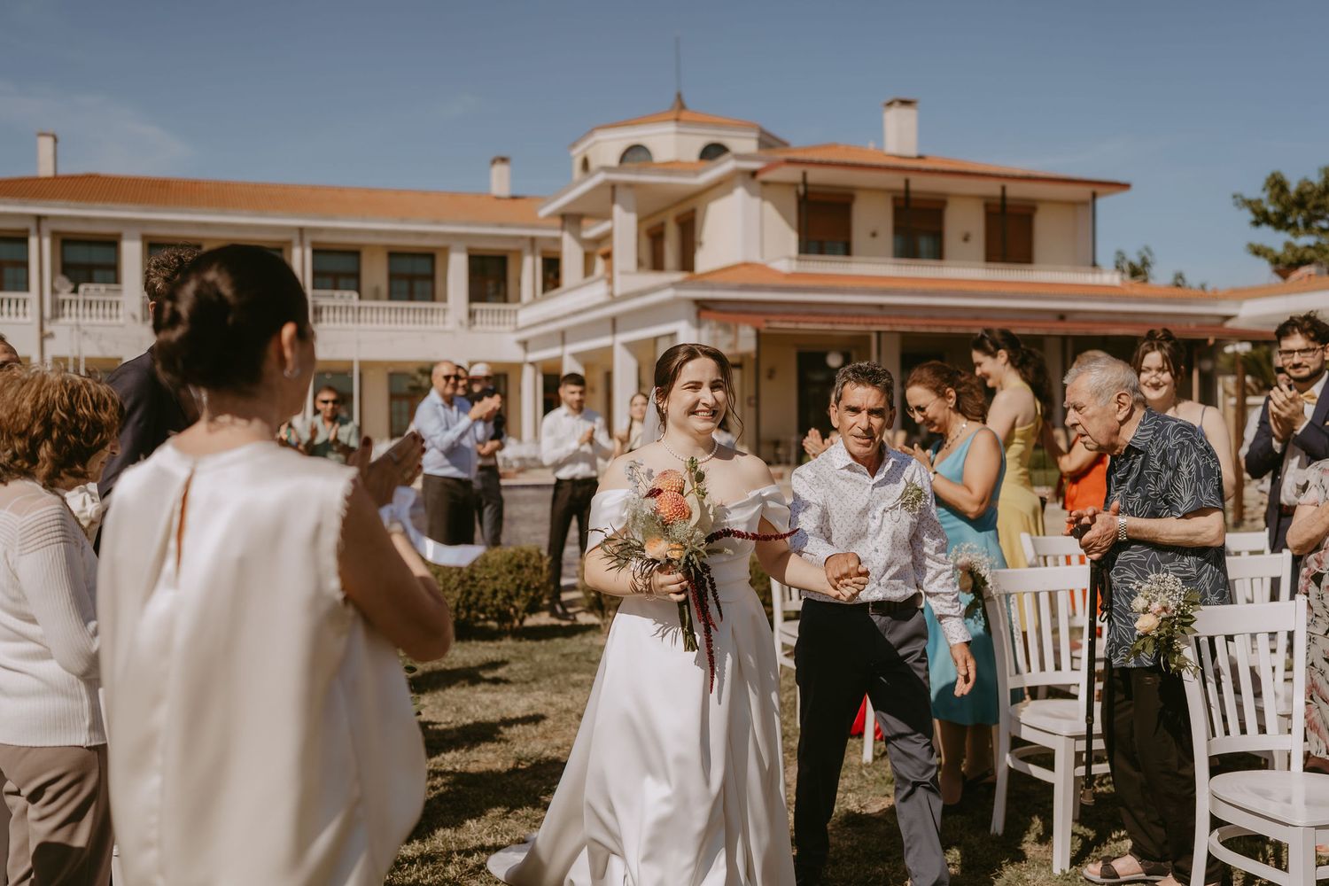 Bride and groom walk down outdoor aisle as guests celebrate at a sunny wedding venue with white chairs.