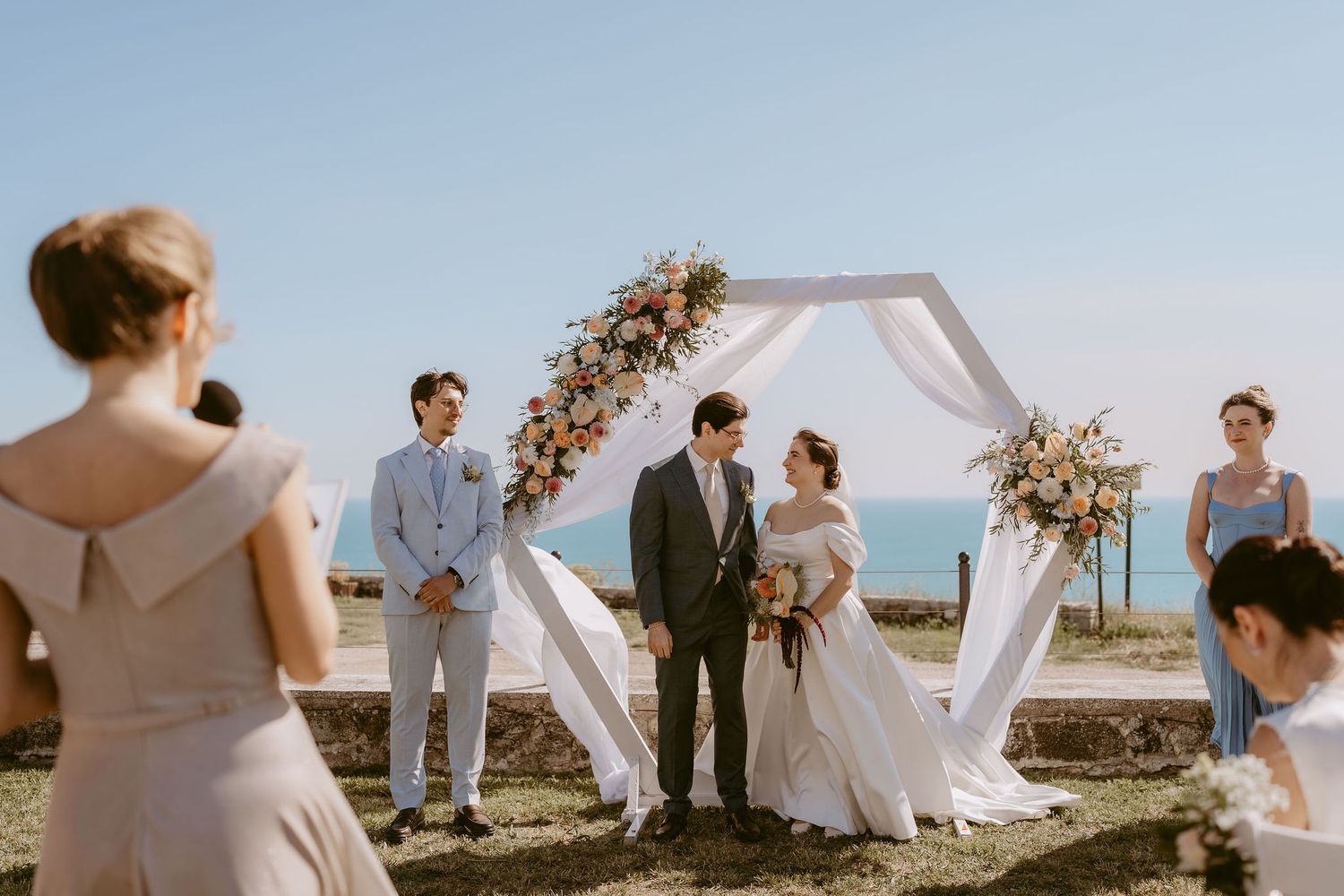 Couple exchanges vows at outdoor wedding ceremony under floral hexagonal arch with ocean view and wedding party.