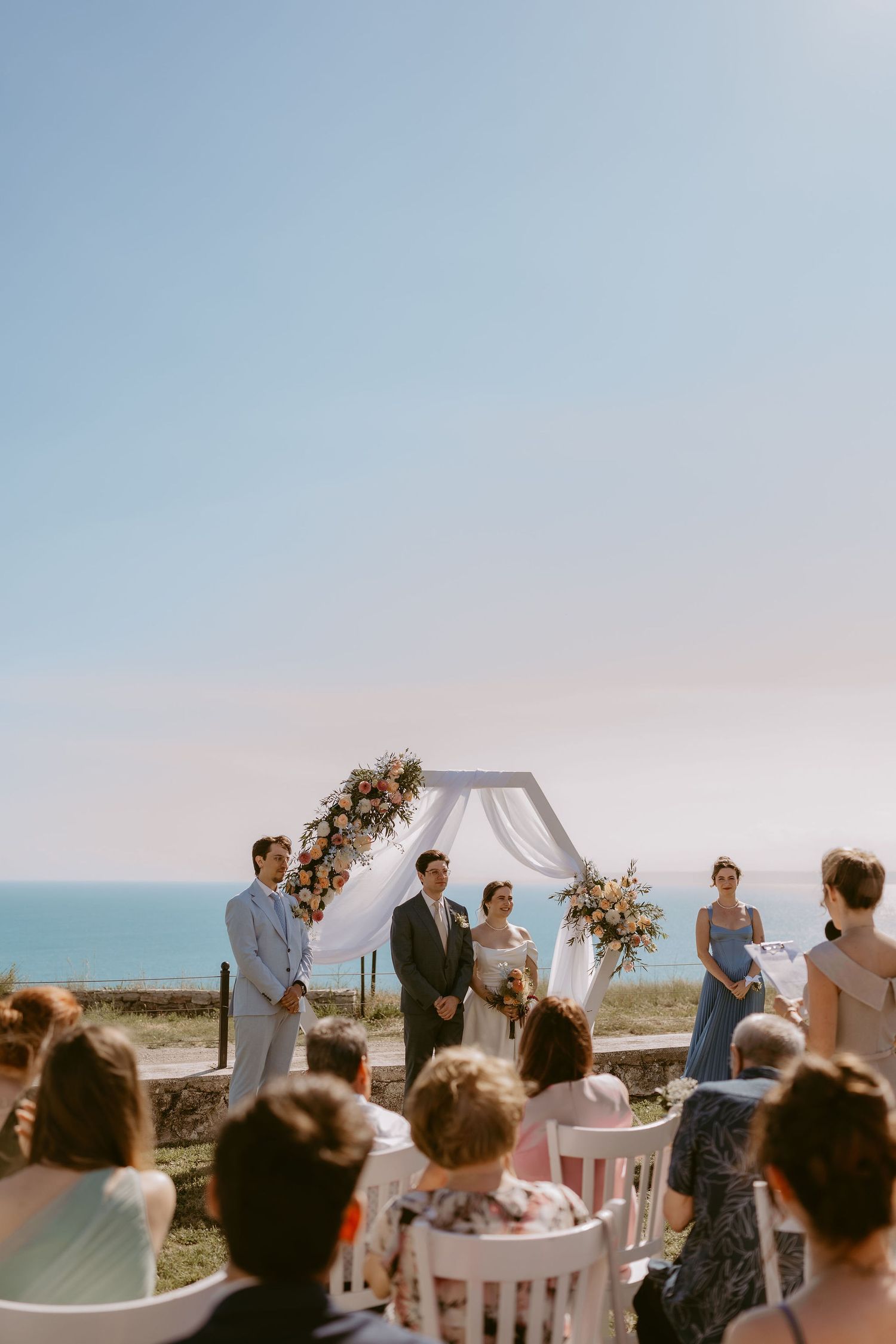 Outdoor beach wedding ceremony with couple at floral arch, guests seated, ocean backdrop on sunny day.
