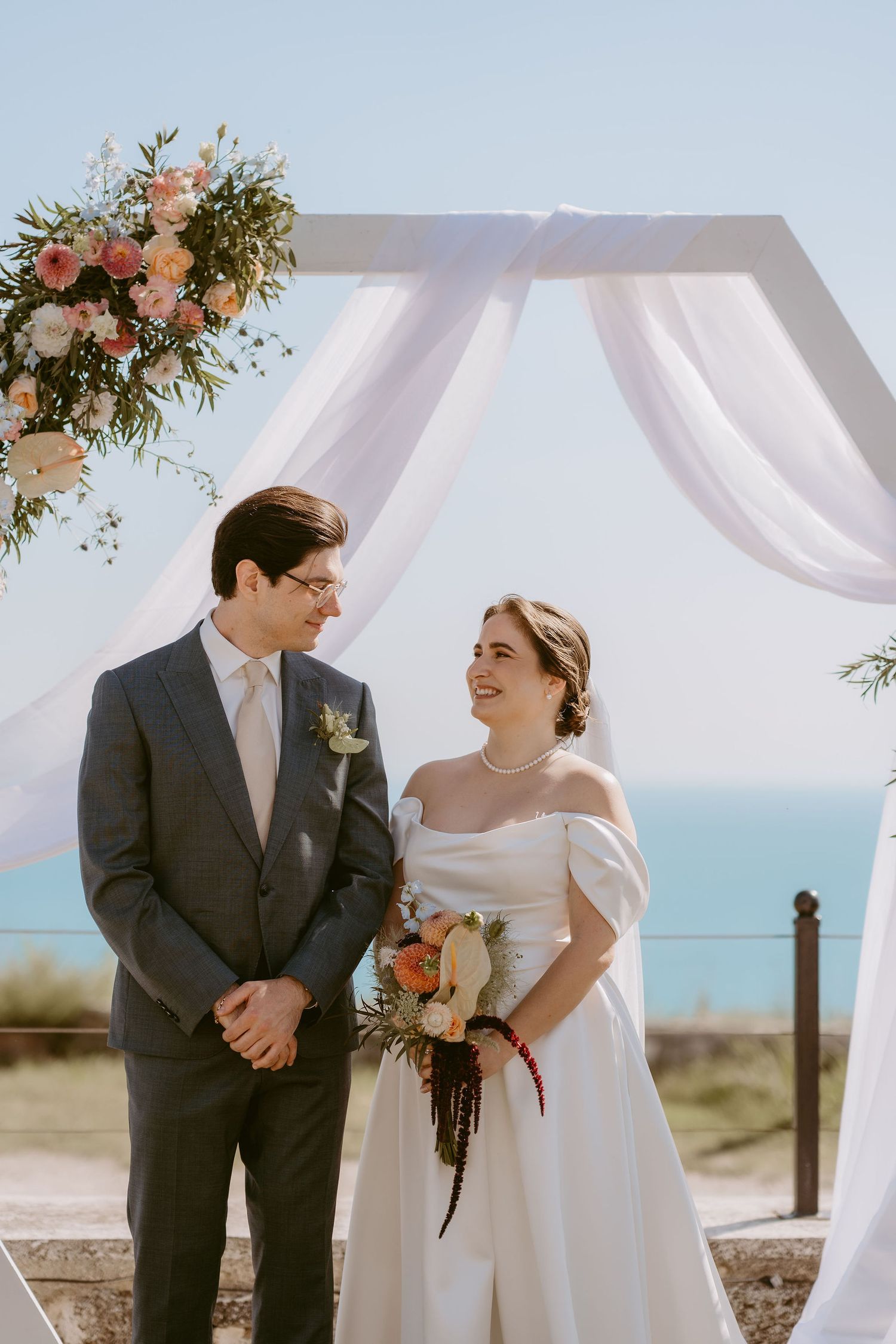 Bride and groom smiling at each other under a floral arch with ocean views on their wedding day.