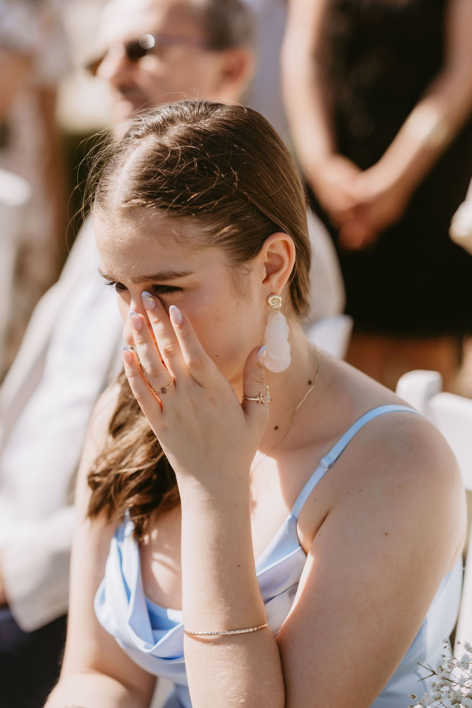 Emotional bridesmaid in blue dress crying and covering face with hand during outdoor wedding ceremony.