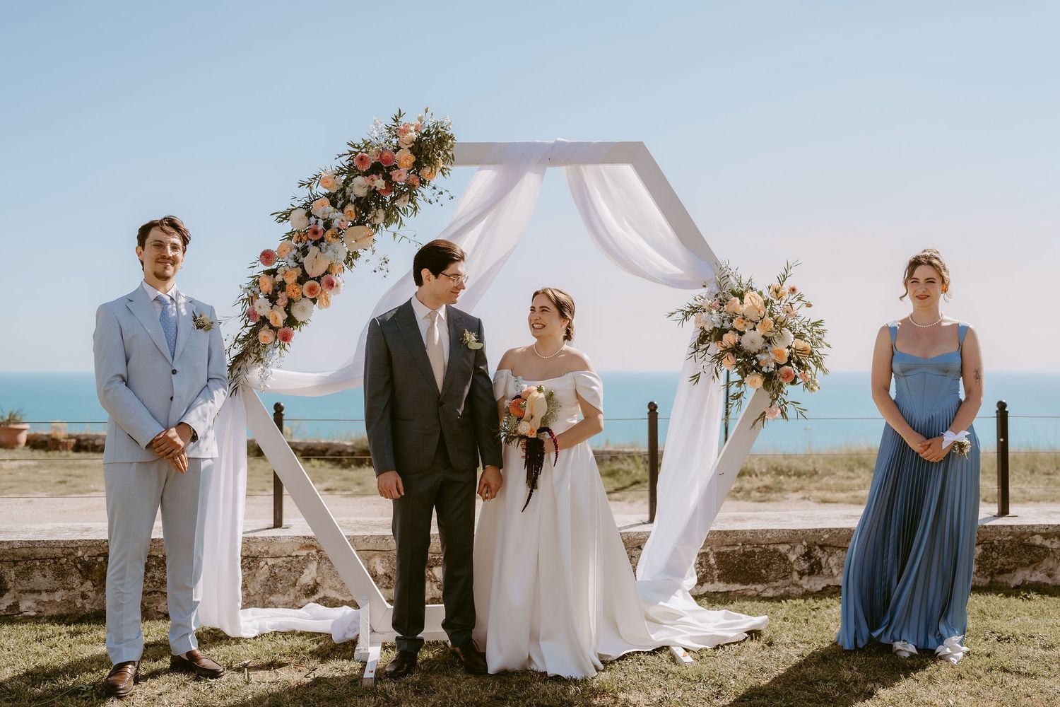 Couple exchanges vows under floral hexagonal arch with ocean backdrop, flanked by groomsman and bridesmaid.
