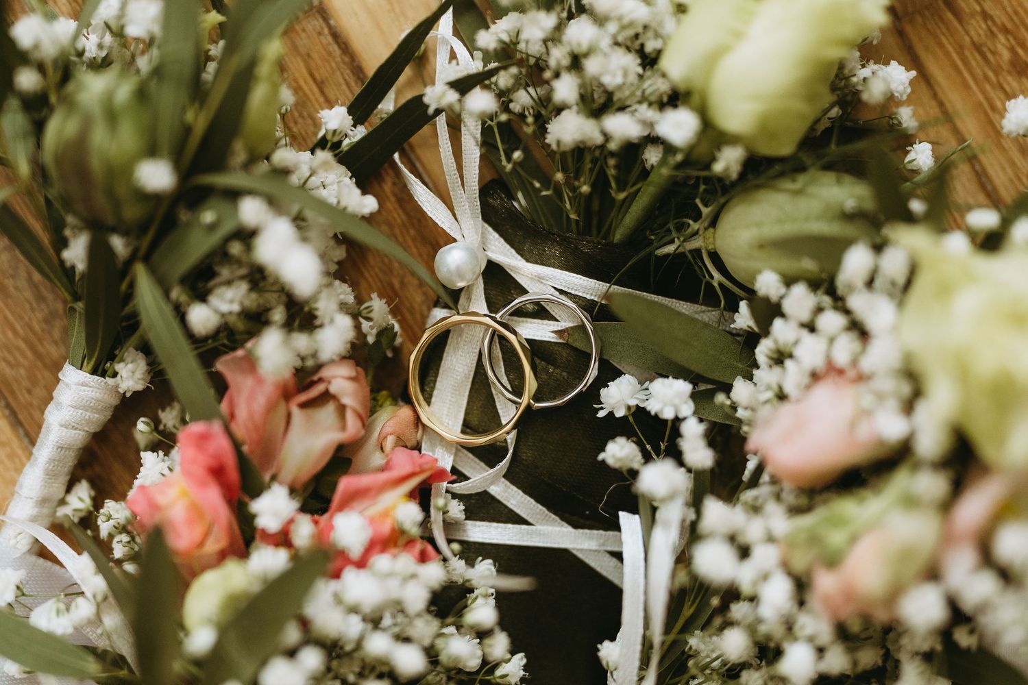Wedding rings resting on white ribbon among baby's breath flowers and pink roses boutonnieres on wooden surface.