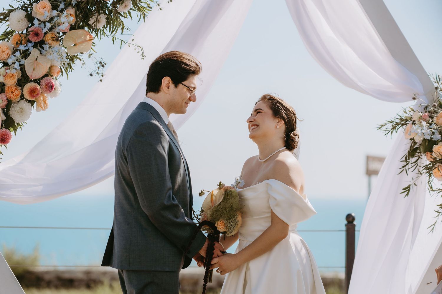Couple exchanging vows at outdoor wedding ceremony under floral arch with ocean view backdrop.