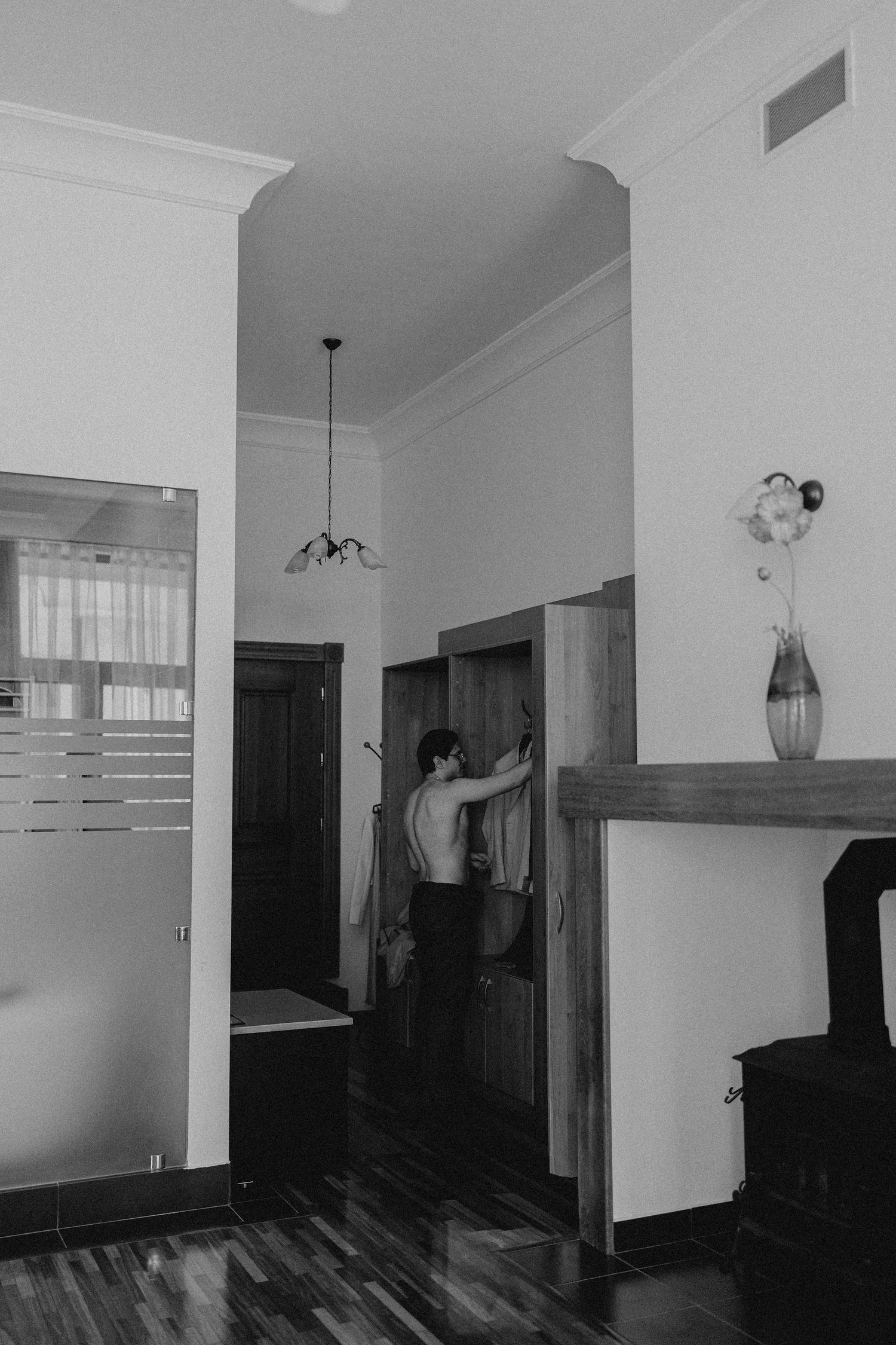 Black and white photo of a shirtless man selecting clothes from a wardrobe in an elegant room with hardwood floors.