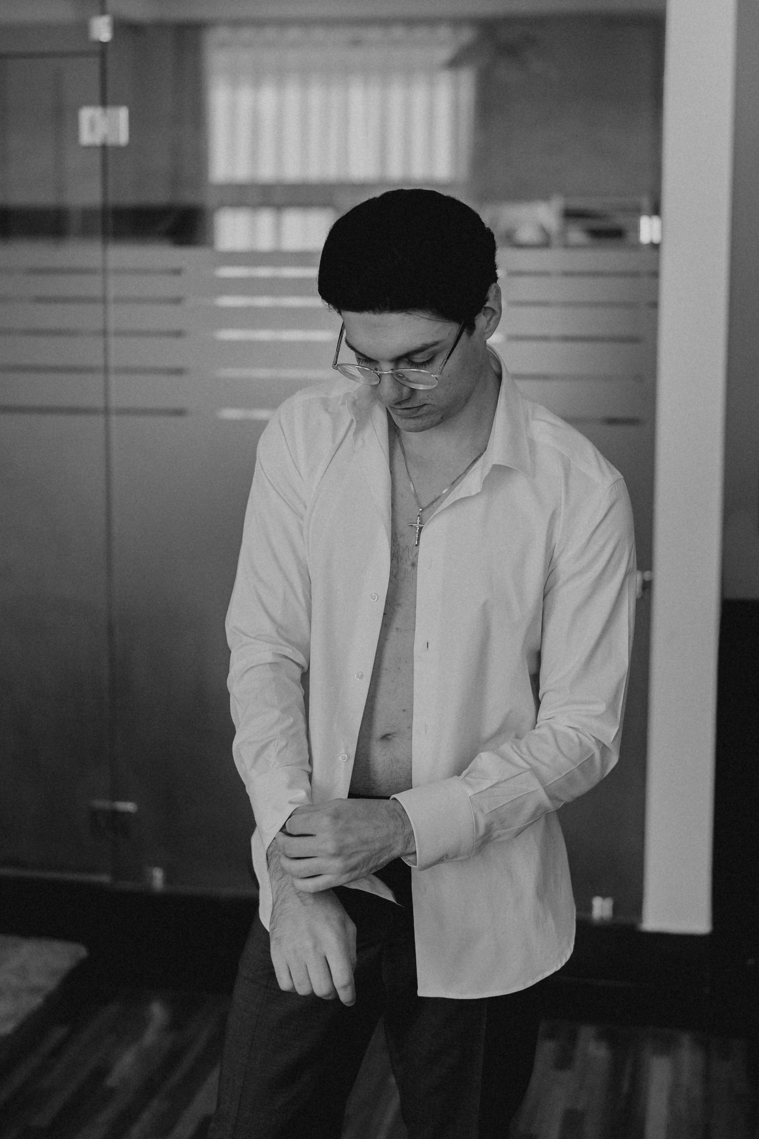 Black and white photo of a man in glasses and beanie buttoning a white dress shirt in a modern office.