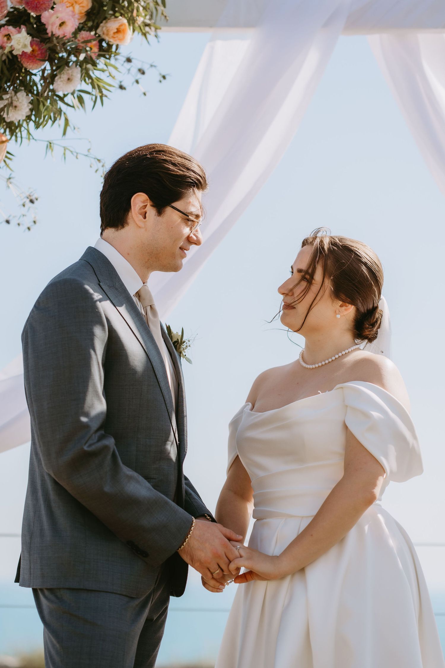 Bride and groom holding hands at outdoor wedding ceremony under floral arch with white draping and blue sky.