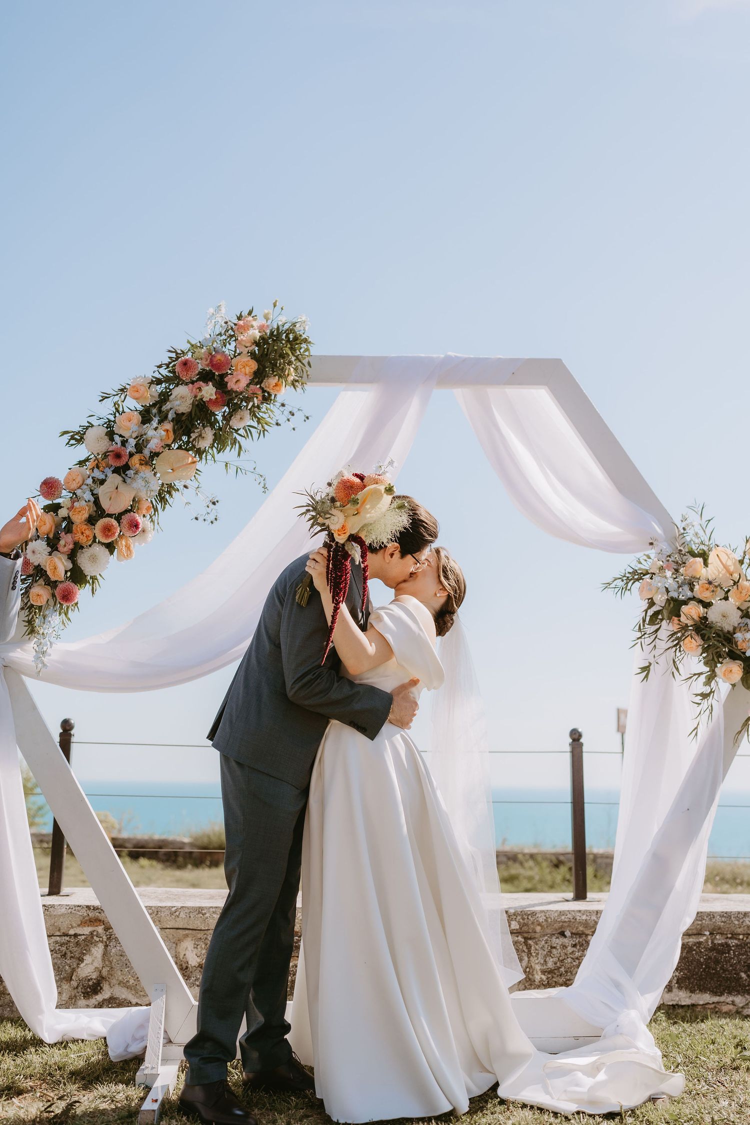 Bride and groom share first kiss under floral hexagonal arch with white draping and ocean view on wedding day.