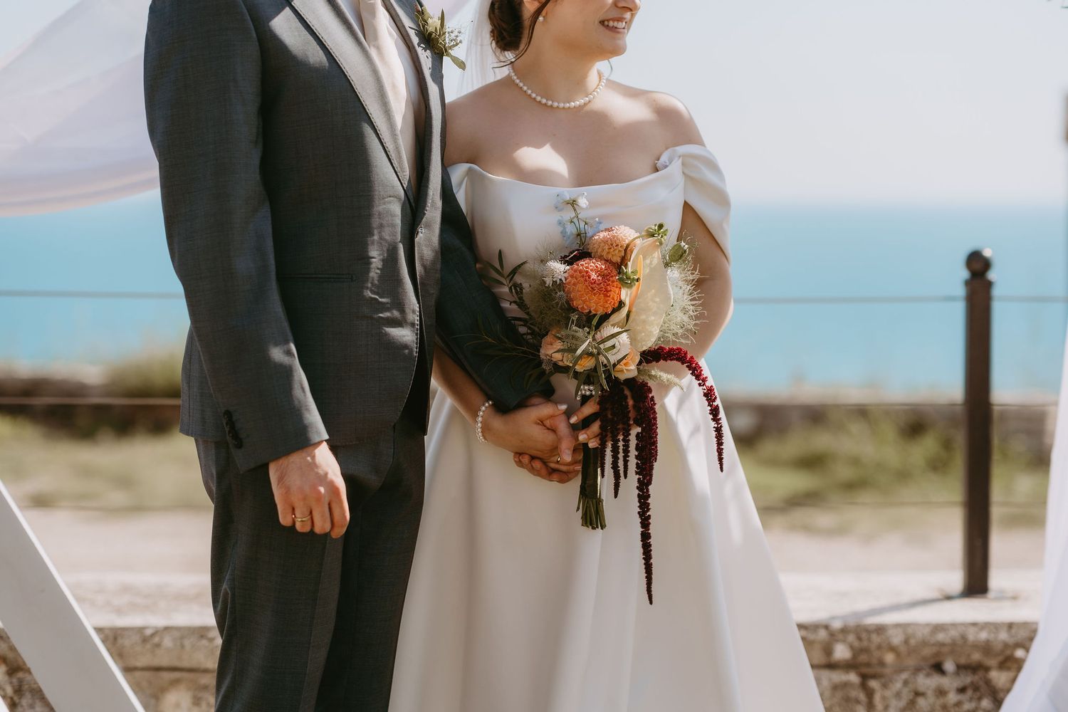 Bride and groom pose together outdoors holding a colorful bouquet with ocean views behind them on their wedding day.