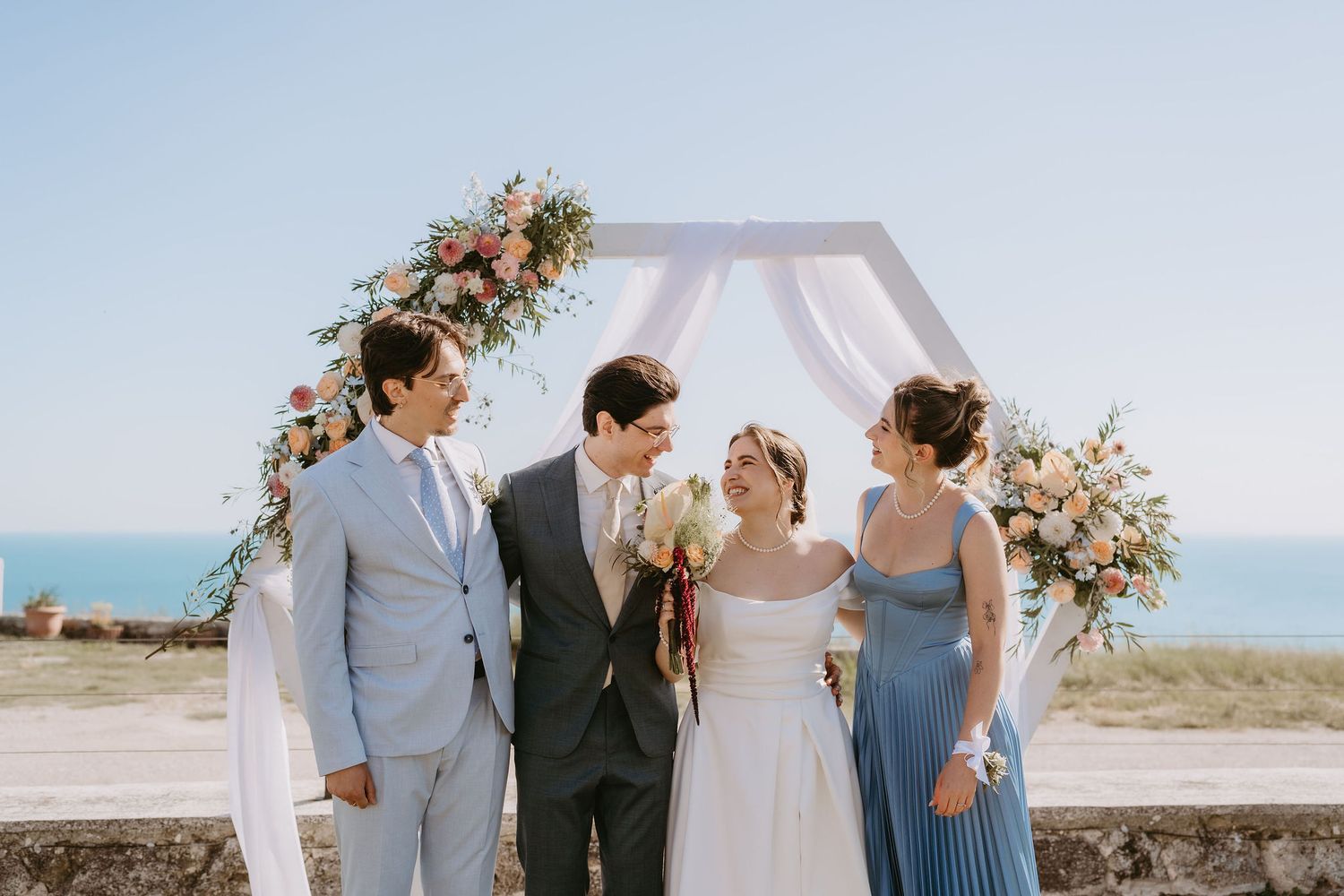 Bride and groom celebrate with two guests under a floral arch with ocean backdrop on a sunny wedding day.