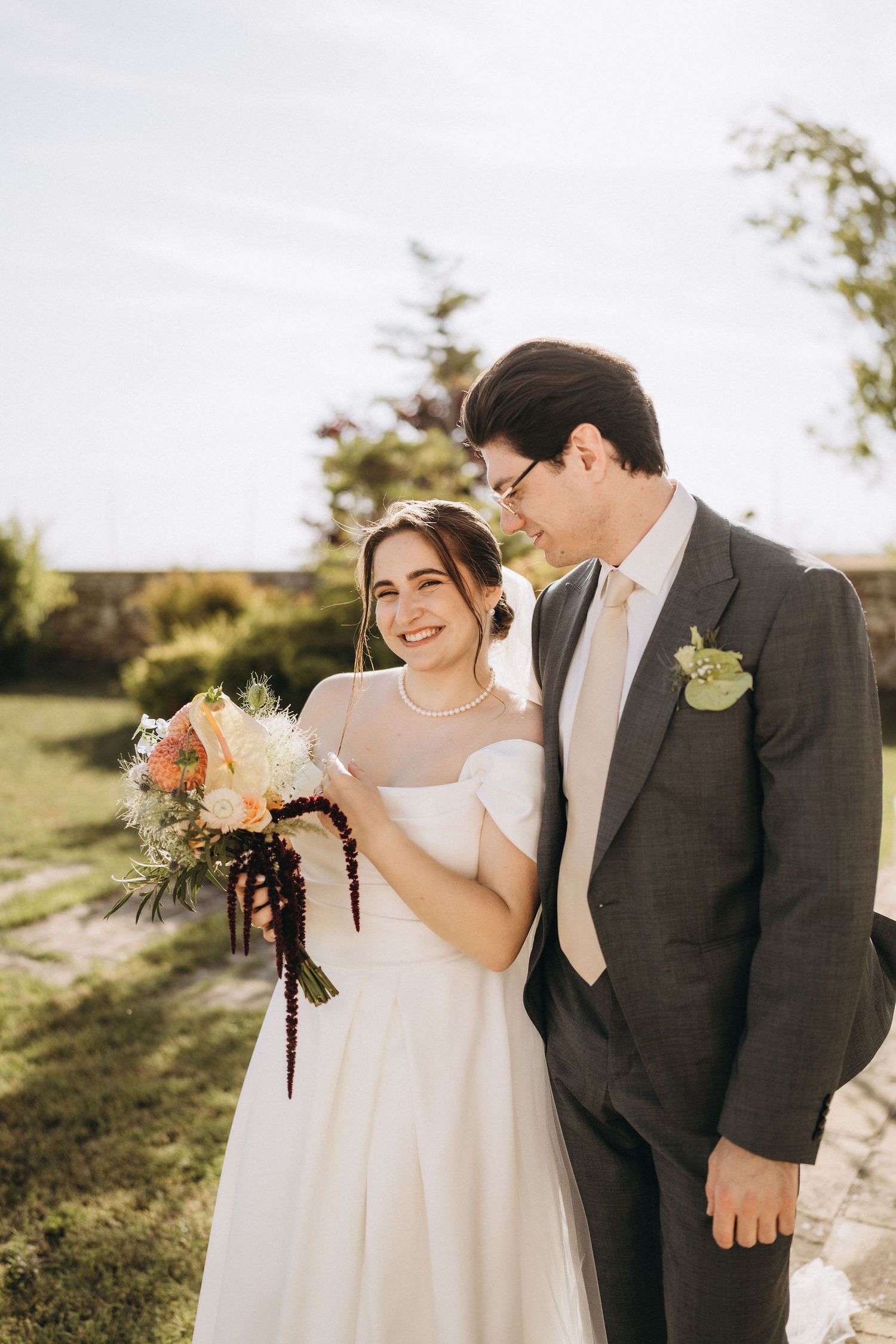 Smiling bride in white dress holding colorful bouquet stands beside groom in gray suit outdoors on sunny wedding day.