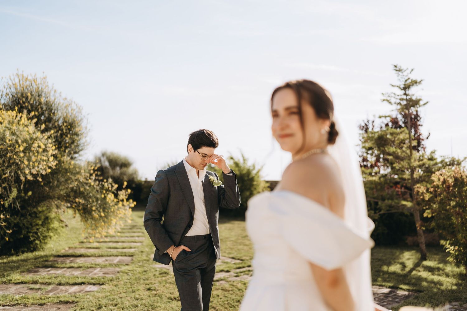 A bride in a white off-shoulder gown stands in the foreground as a groom in a gray suit walks along a garden path.