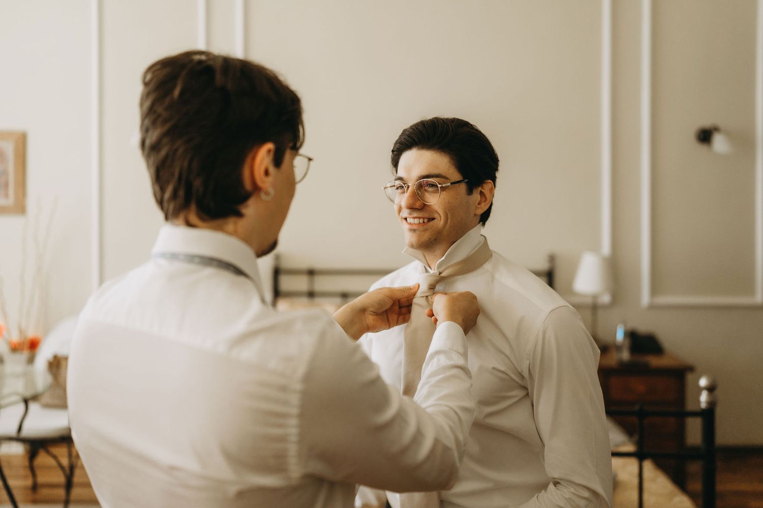 Two men in white dress shirts, one adjusting the other's collar and tie while smiling indoors.