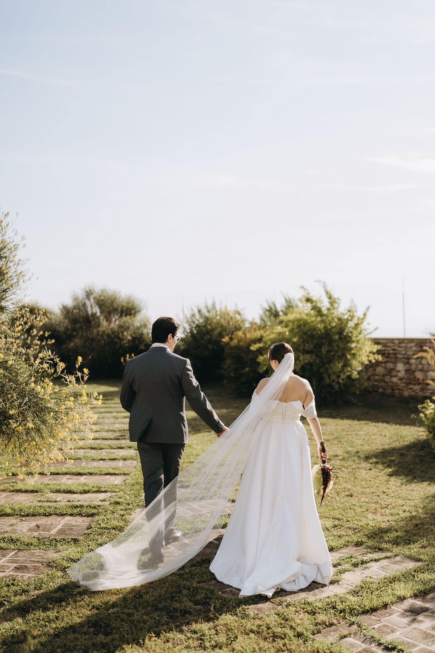 Bride and groom walking hand in hand through a lush garden, bride wearing a flowing white gown with long veil.