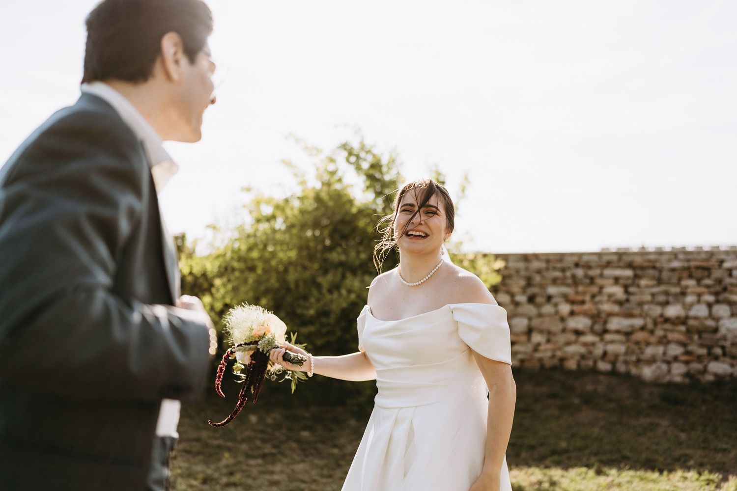 Bride in off-shoulder white dress laughs joyfully holding bouquet outdoors near stone wall on sunny wedding day.