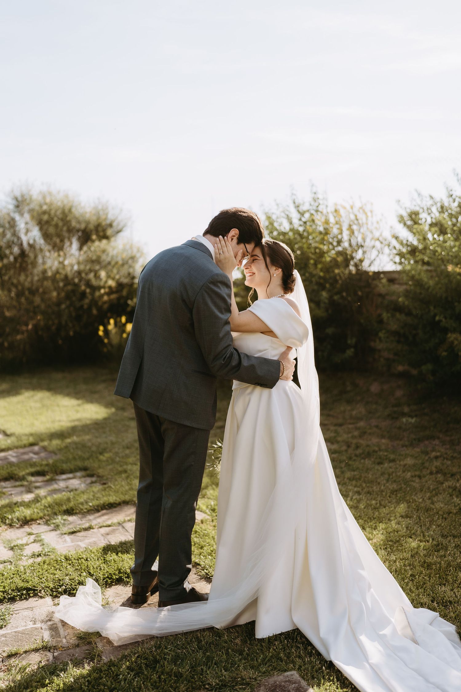 Bride and groom share a romantic moment in garden, she in elegant white gown with train, he in gray suit.