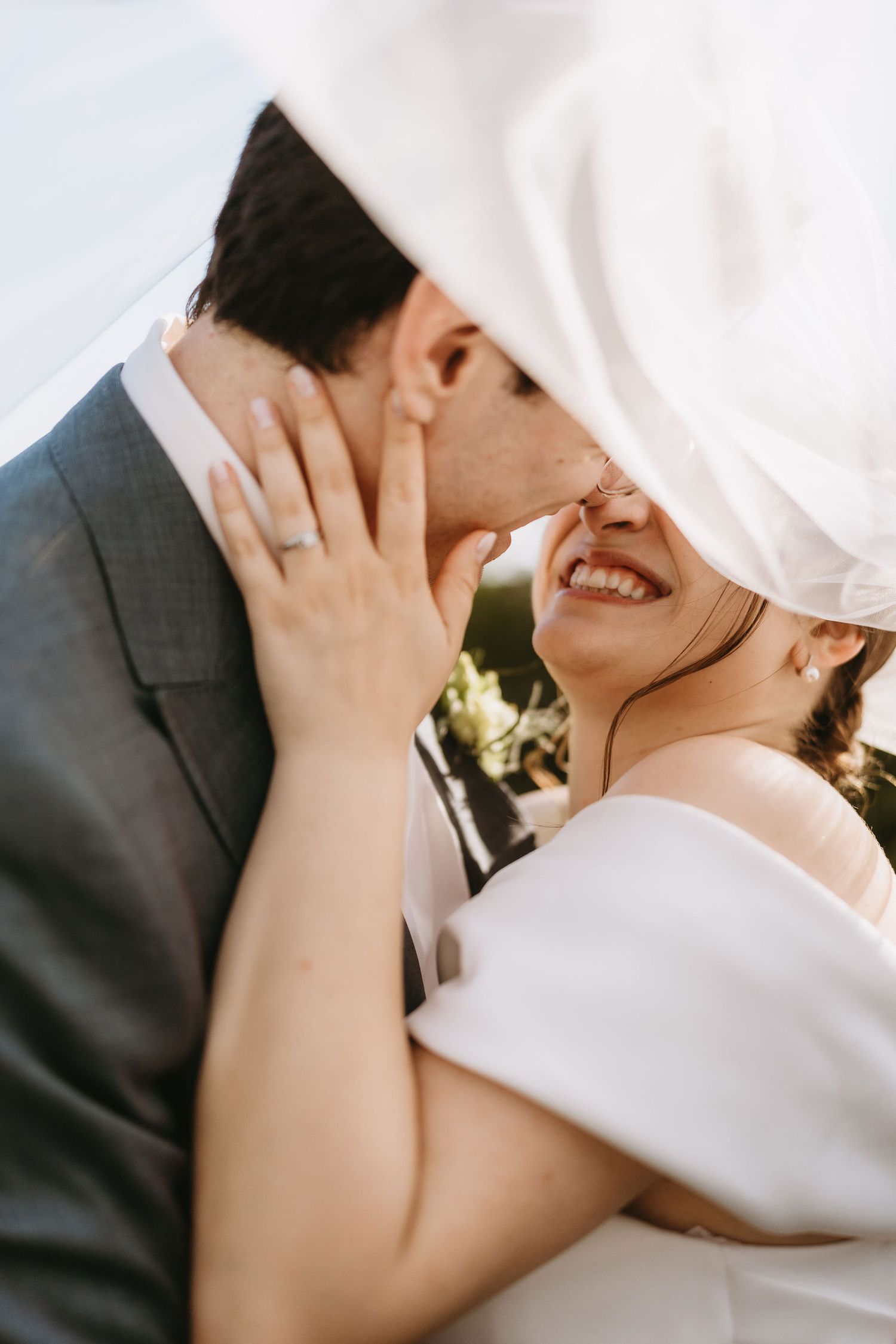 Bride and groom sharing a romantic moment under a white veil, bride smiling with wedding ring visible.