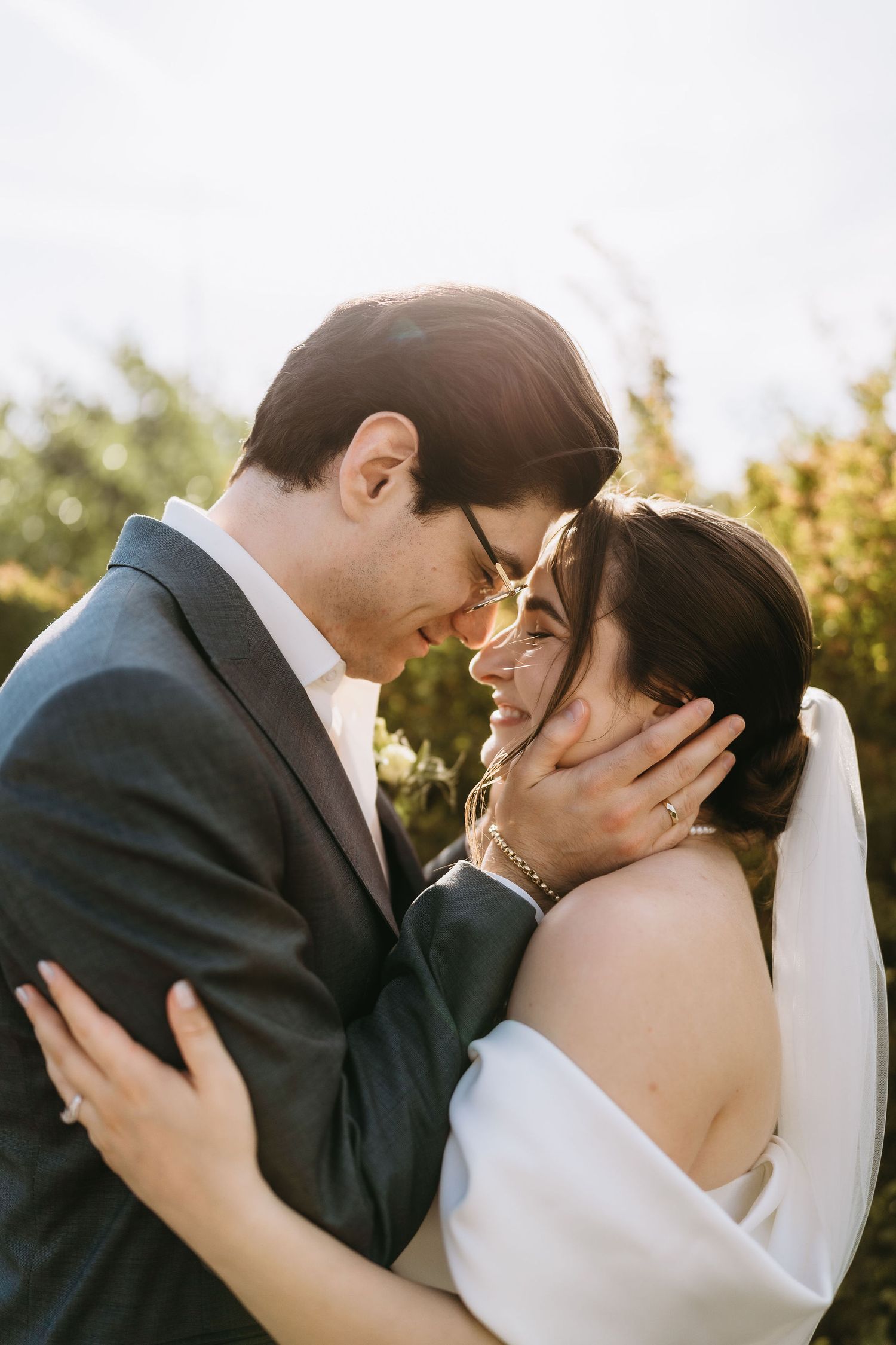 A bride and groom share a romantic forehead touch outdoors, bathed in golden sunlight on their wedding day.