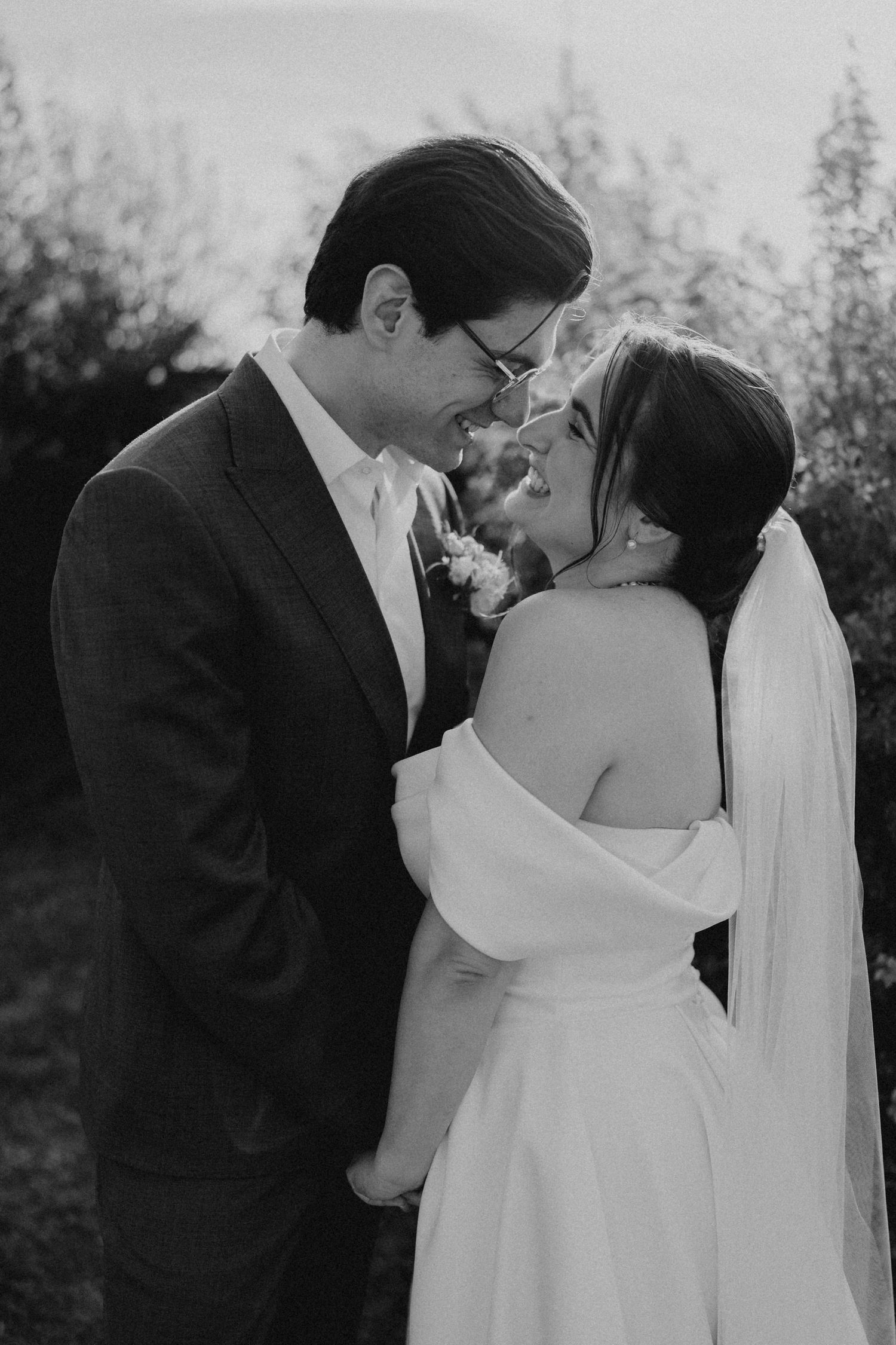 Black and white photo of a smiling bride and groom in wedding attire sharing an intimate moment outdoors.
