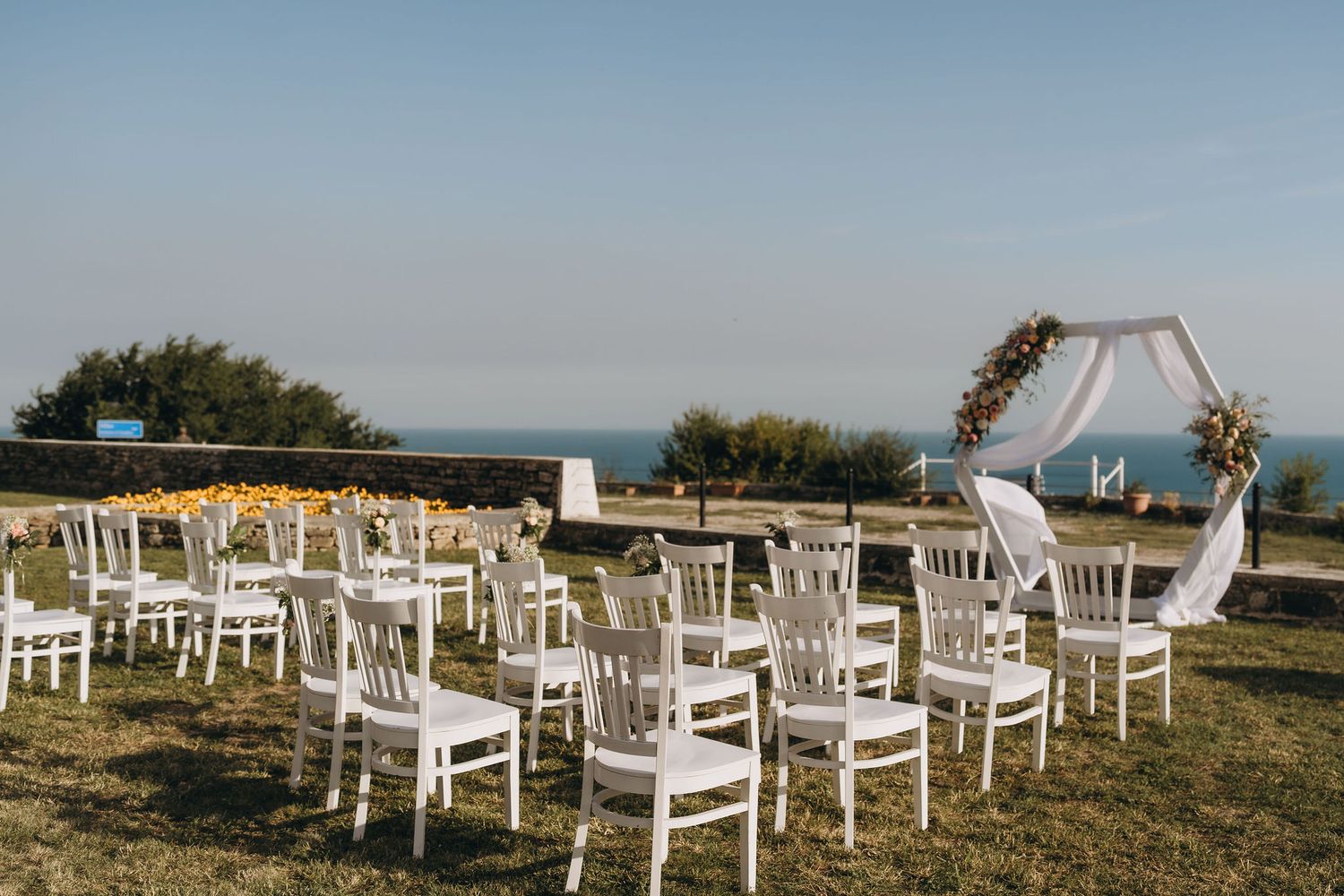 Outdoor wedding ceremony setup with white chairs and floral arch overlooking the ocean on a sunny day.