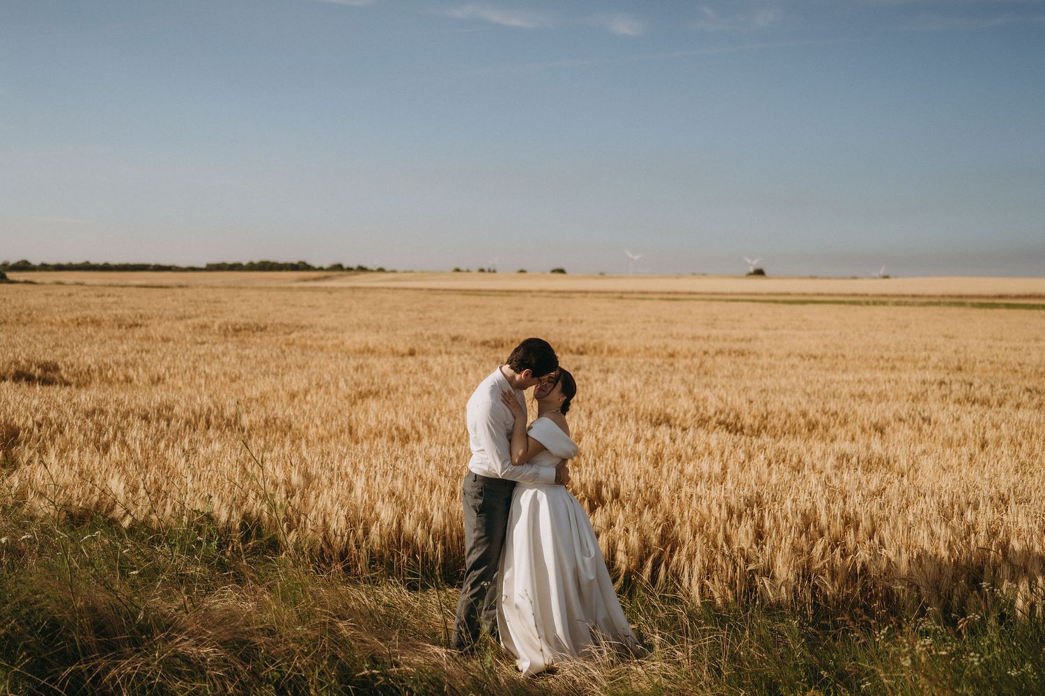 Bride and groom embracing in a golden wheat field under a clear blue sky on their wedding day.