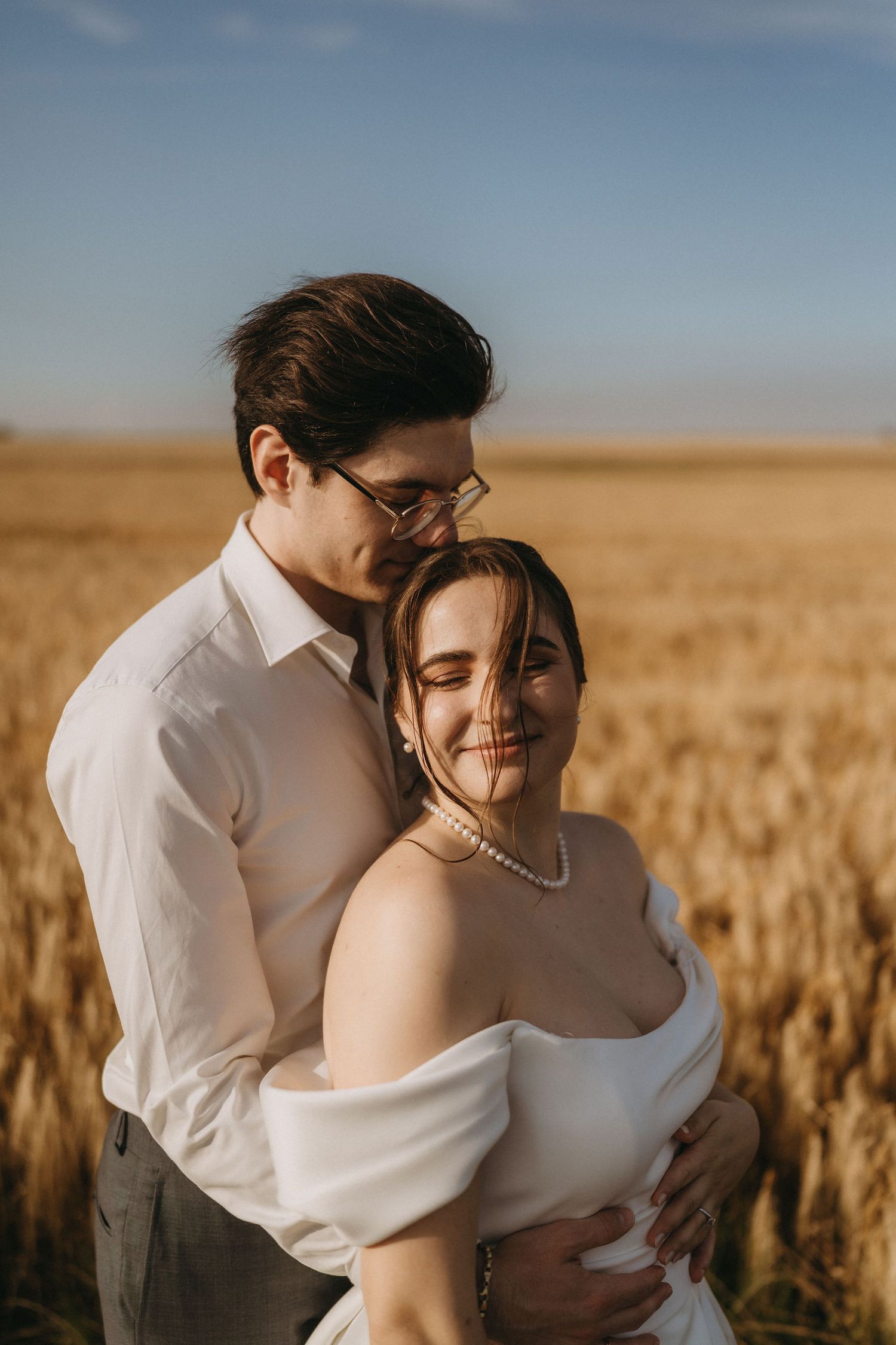 Couple embracing in golden wheat field, bride in white off-shoulder gown with pearl necklace, groom in white shirt and glasses.