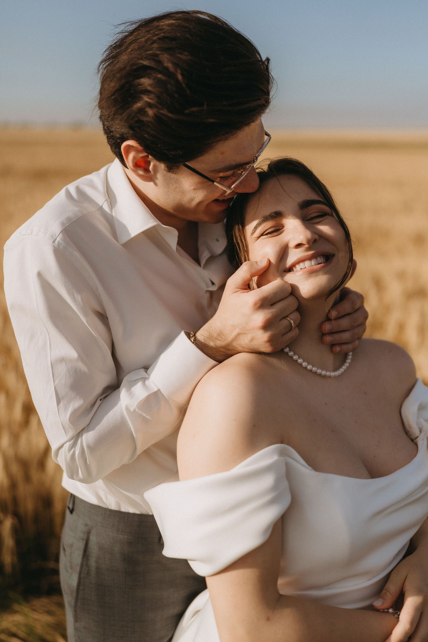 A groom in glasses kisses his smiling bride in a white off-shoulder gown in a golden wheat field.