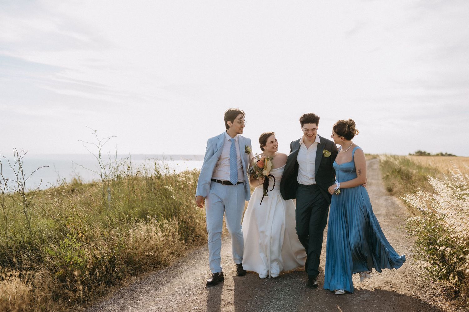 Wedding party of four walking on coastal path, bride in white gown, bridesmaids in blue dresses, groomsmen in suits.