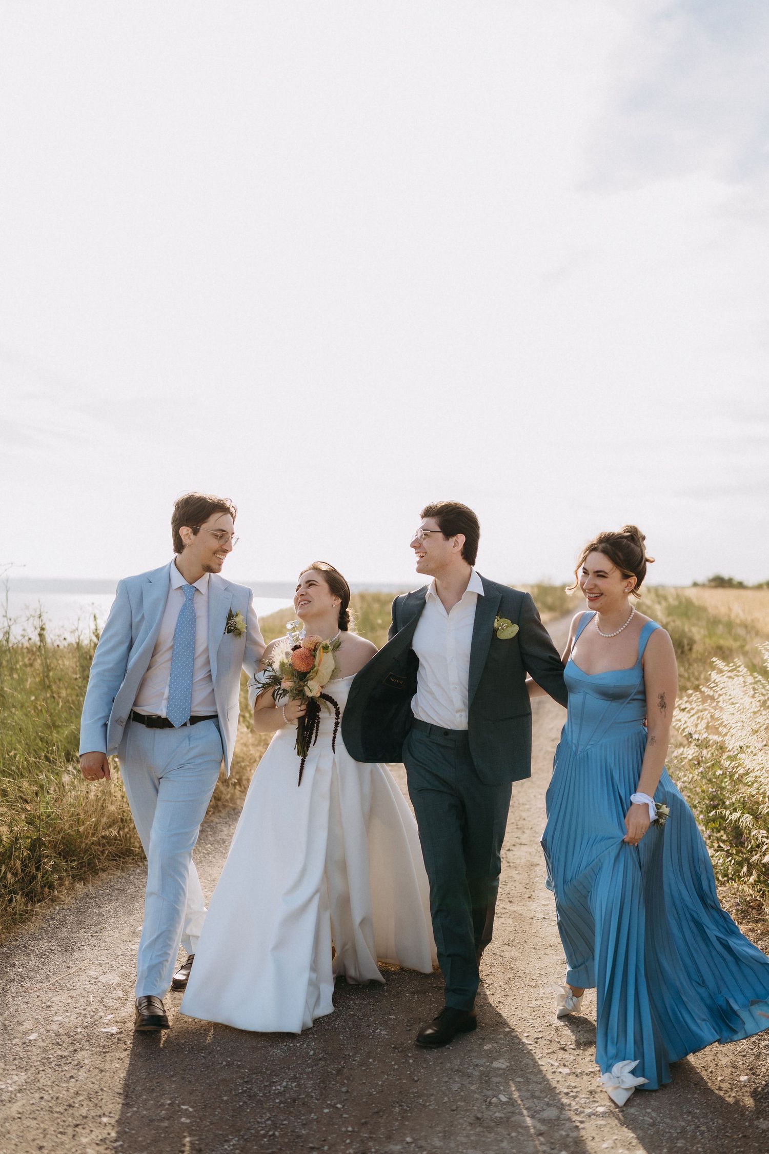 Wedding party walking outdoors, bride in white gown with bouquet, groom in navy suit, groomsman in blue, bridesmaid in blue gown.