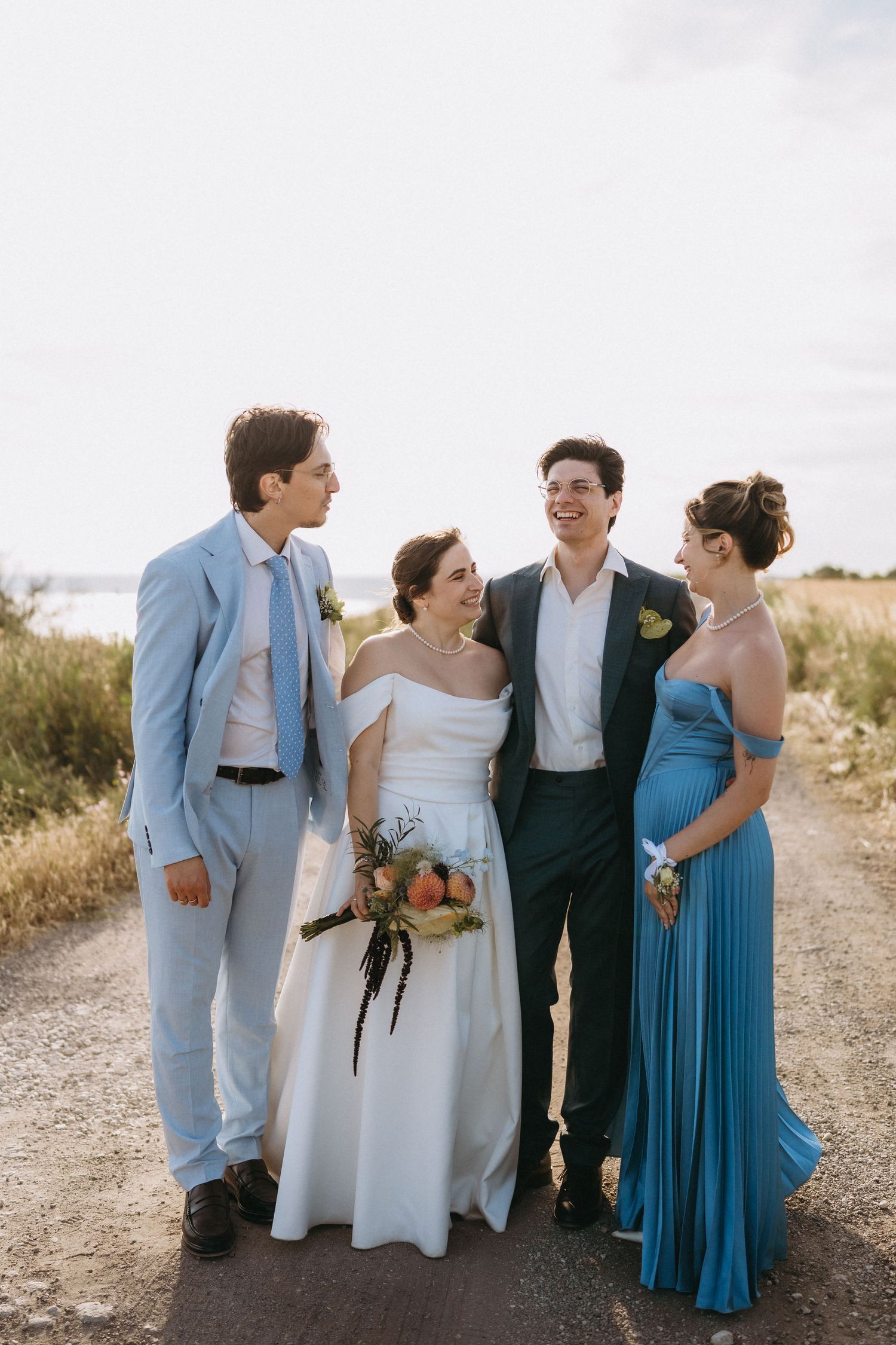 Wedding party of four posing on a dirt path, bride in white gown with bouquet, groom in dark suit, groomsman and bridesmaid in blue.