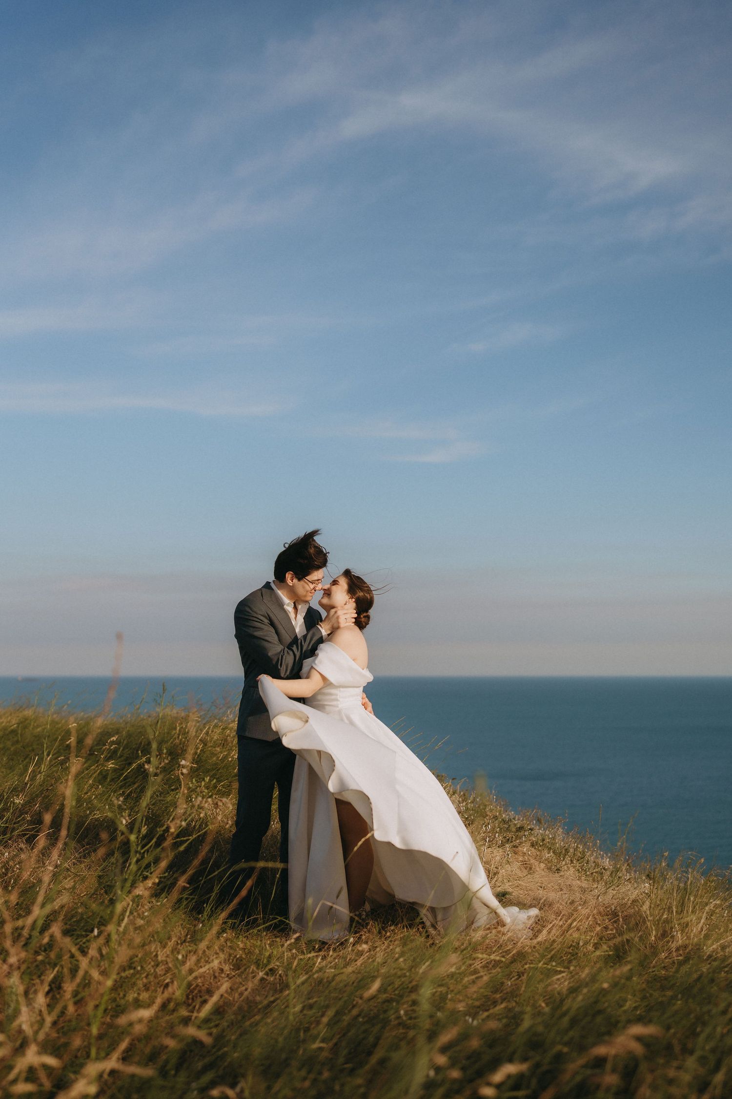 A couple shares a romantic kiss on a coastal cliff, bride in flowing white gown, groom in gray suit, ocean backdrop.