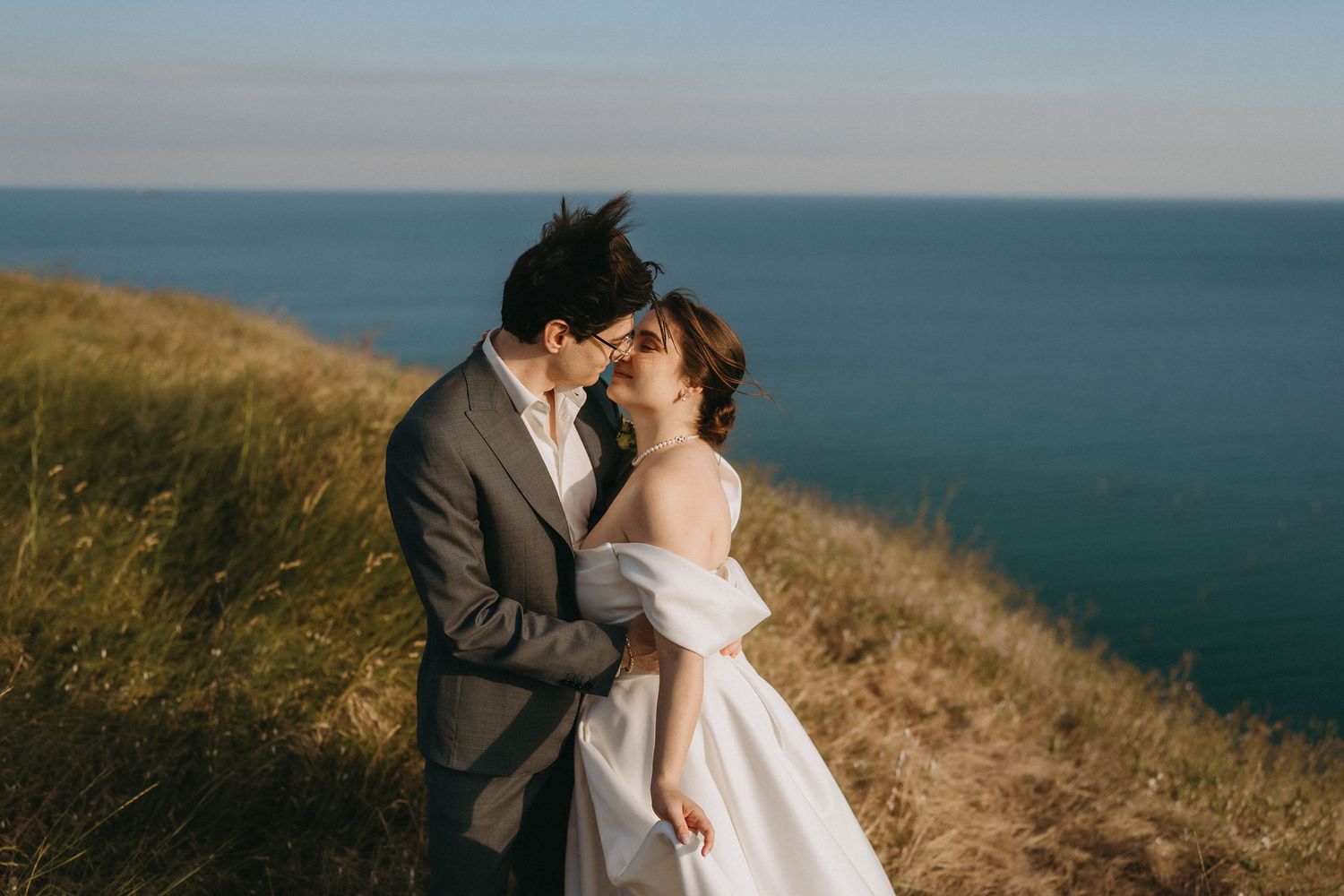 A couple shares a romantic kiss on a coastal cliff, bride in off-shoulder gown, groom in gray suit, ocean backdrop.