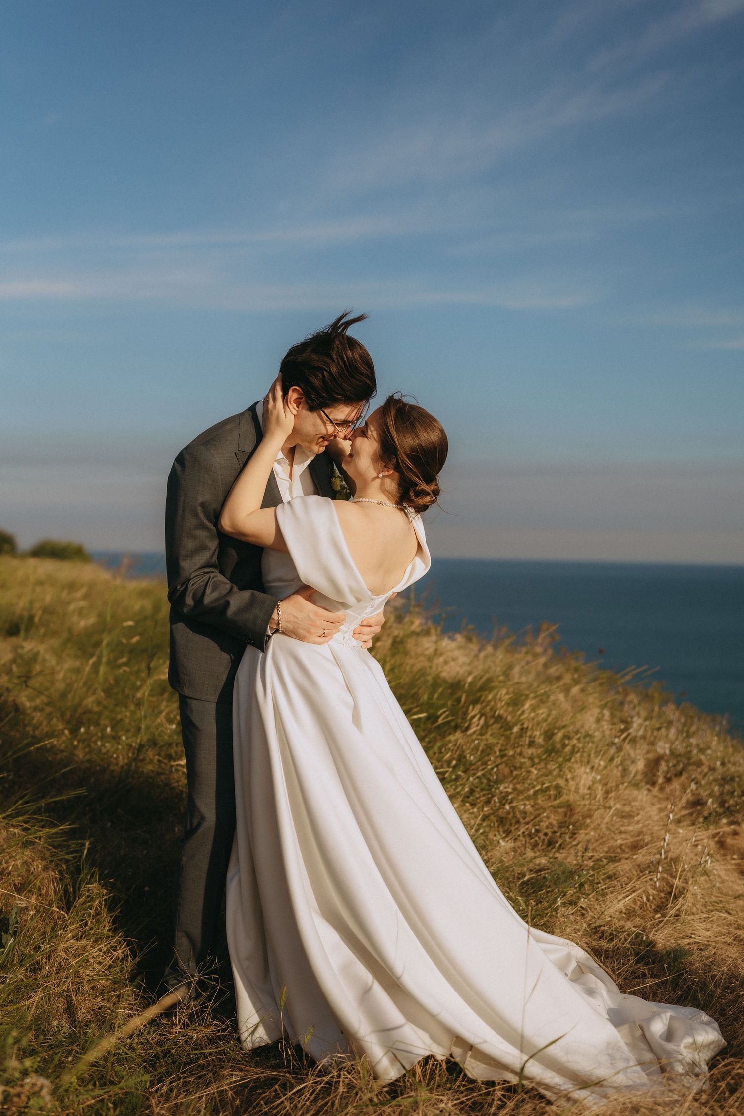 A bride and groom share a romantic kiss on a coastal cliff, surrounded by golden grass and a blue ocean backdrop.