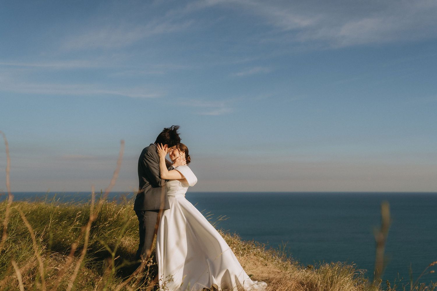 Couple embracing in wedding attire on coastal cliffside with ocean and blue sky backdrop.