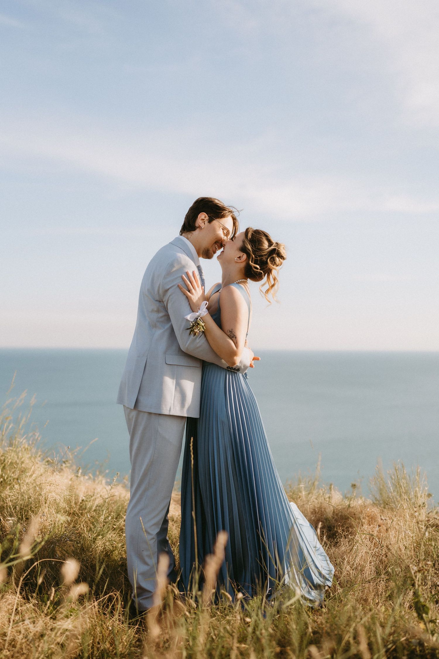 Couple embracing on a coastal cliff, man in gray suit, woman in blue pleated dress, with ocean backdrop.