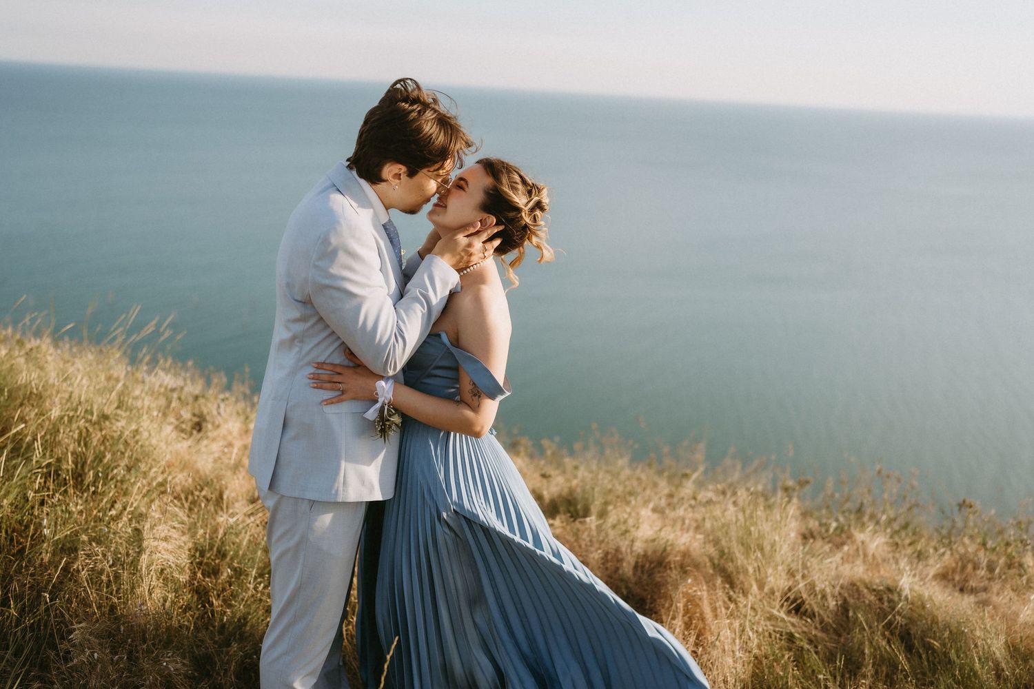 Romantic couple sharing a kiss on a coastal cliff, woman in blue pleated gown, man in white suit, ocean backdrop.