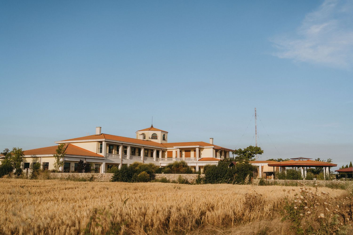 Large Spanish-style villa with terracotta roof surrounded by golden wheat fields under a clear blue sky.
