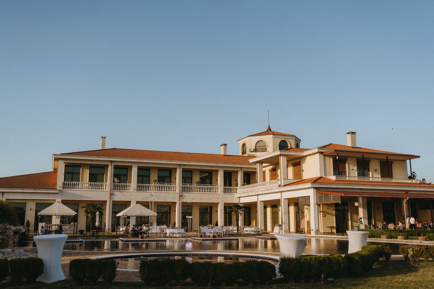 Elegant two-story villa with terracotta roof, outdoor pool, and manicured gardens under a clear blue sky.