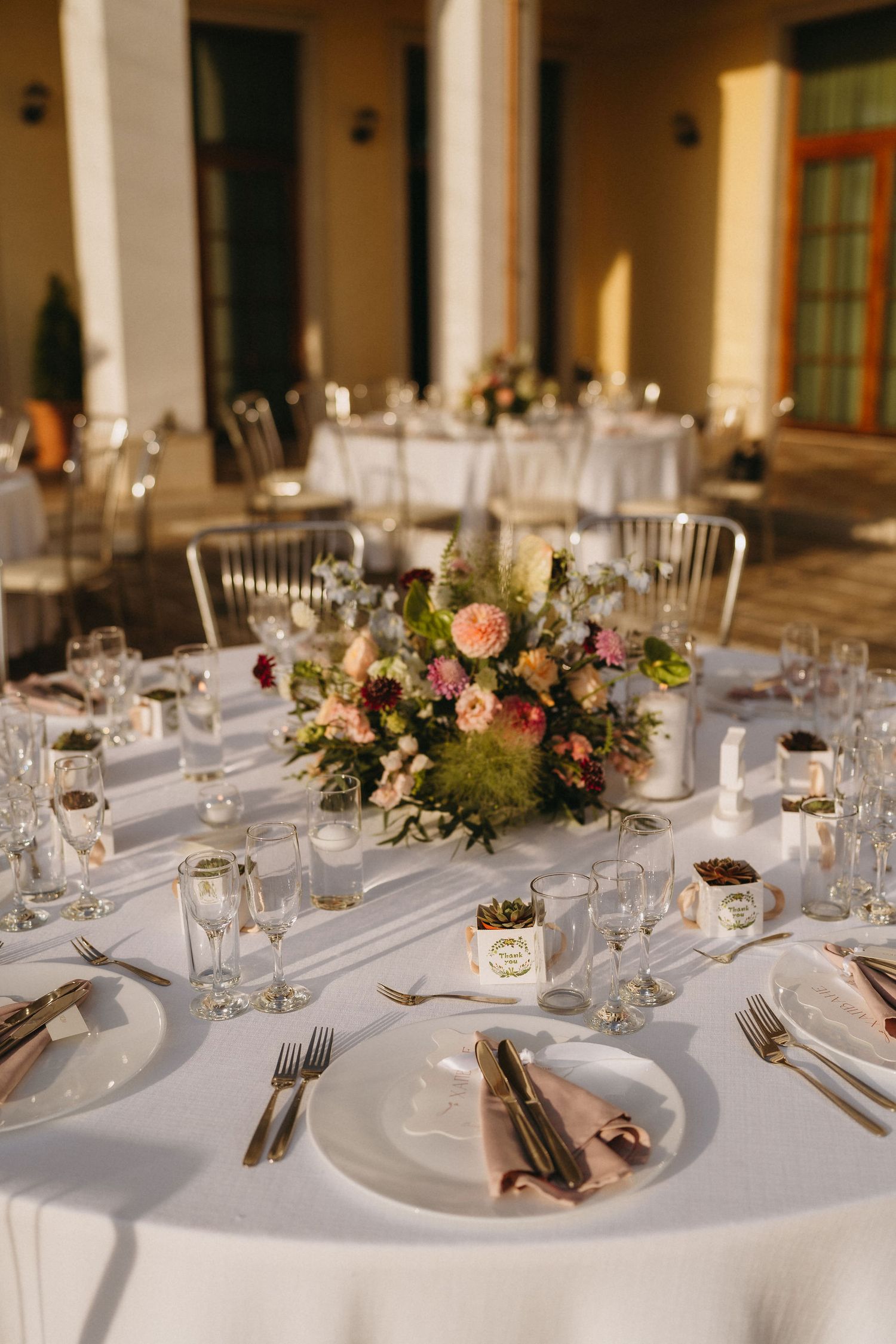 Elegant wedding reception table with white linen, floral centerpiece, crystal glasses, and pink napkins set outdoors.