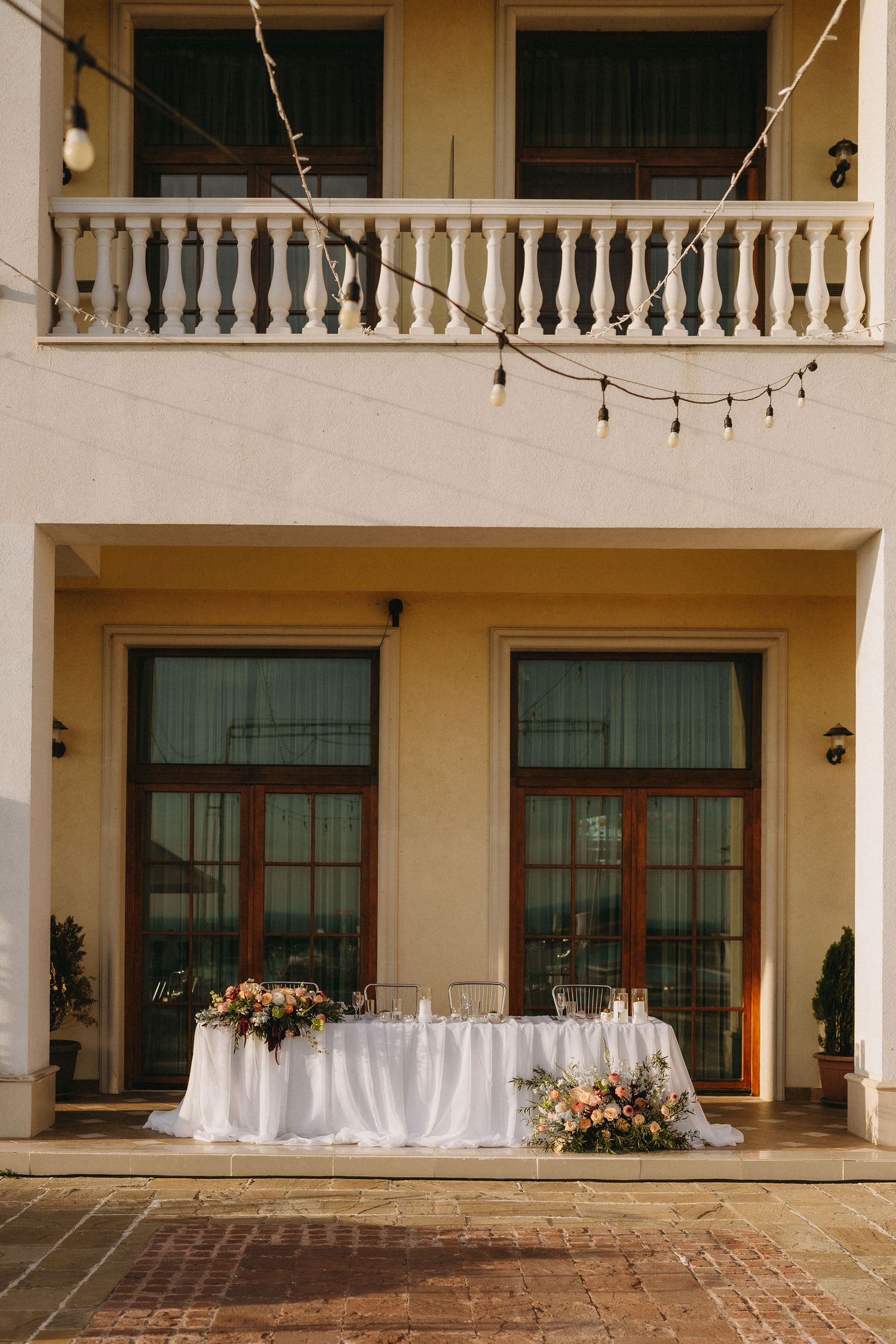Elegant outdoor wedding reception sweetheart table with white linens and floral arrangements against a classic building facade.