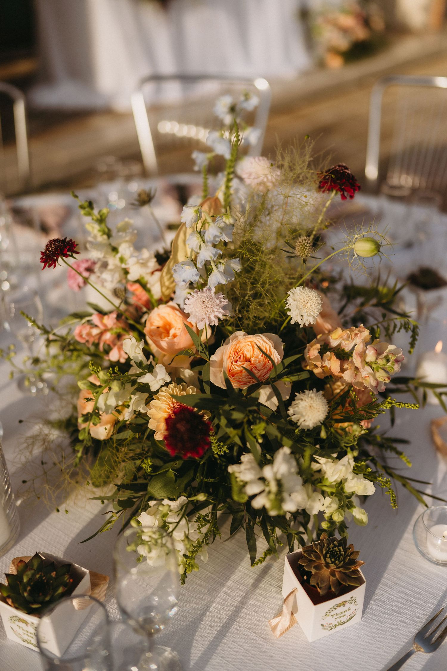 Elegant wedding centerpiece with peach roses, wildflowers, and greenery on a white tablecloth with succulent favors.
