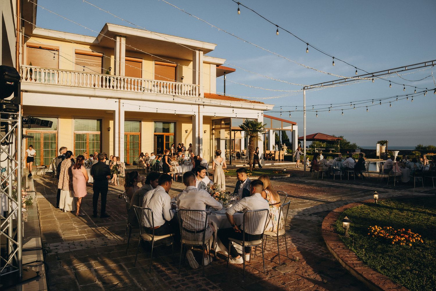 Outdoor evening reception at elegant venue with string lights, guests dining on patio near two-story building at golden hour.